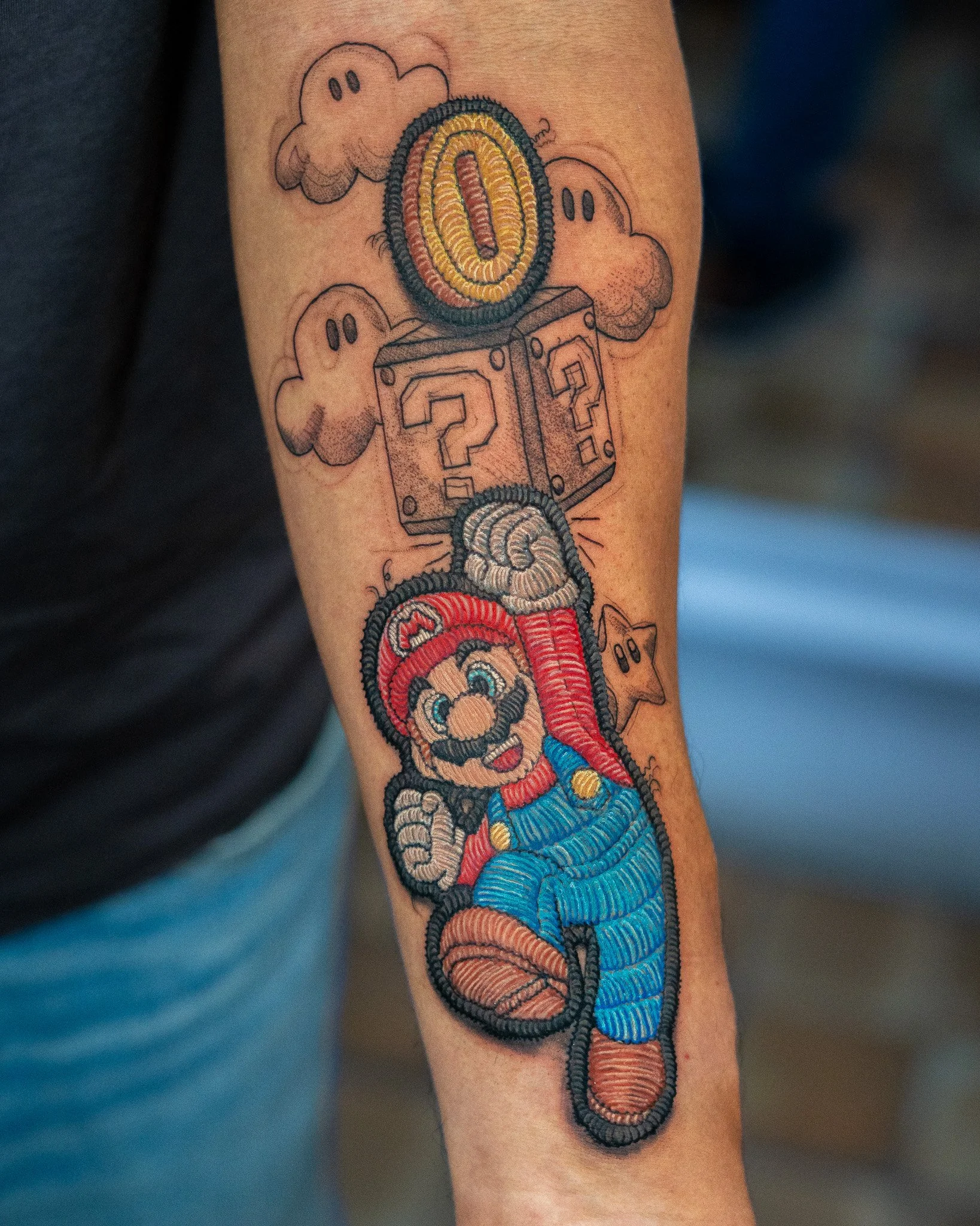Tatuaje de estilo de Super Mario en el brazo, que muestra una serie de bloques con signos de interrogación y monedas, con nubes alrededor.  tatuaje estilo parche hecho en Antigua Guatemala, Guatemala. Tatuaje tejido, tatuaje con efecto de hilos