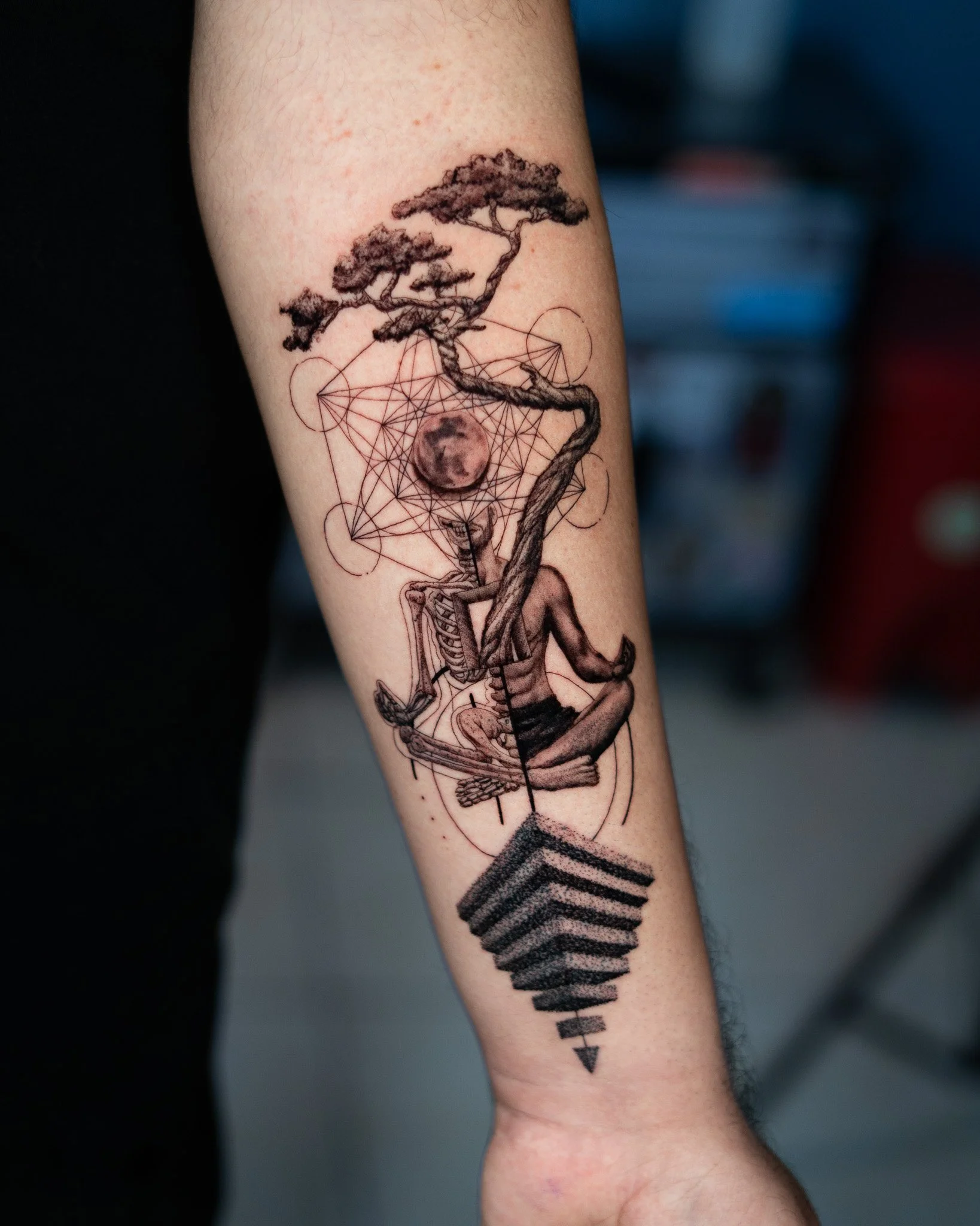 Tatuaje en el brazo de una persona que muestra un árbol de estilo japonés sobre un fondo de líneas geométricas y elementos geométricos adicionales.