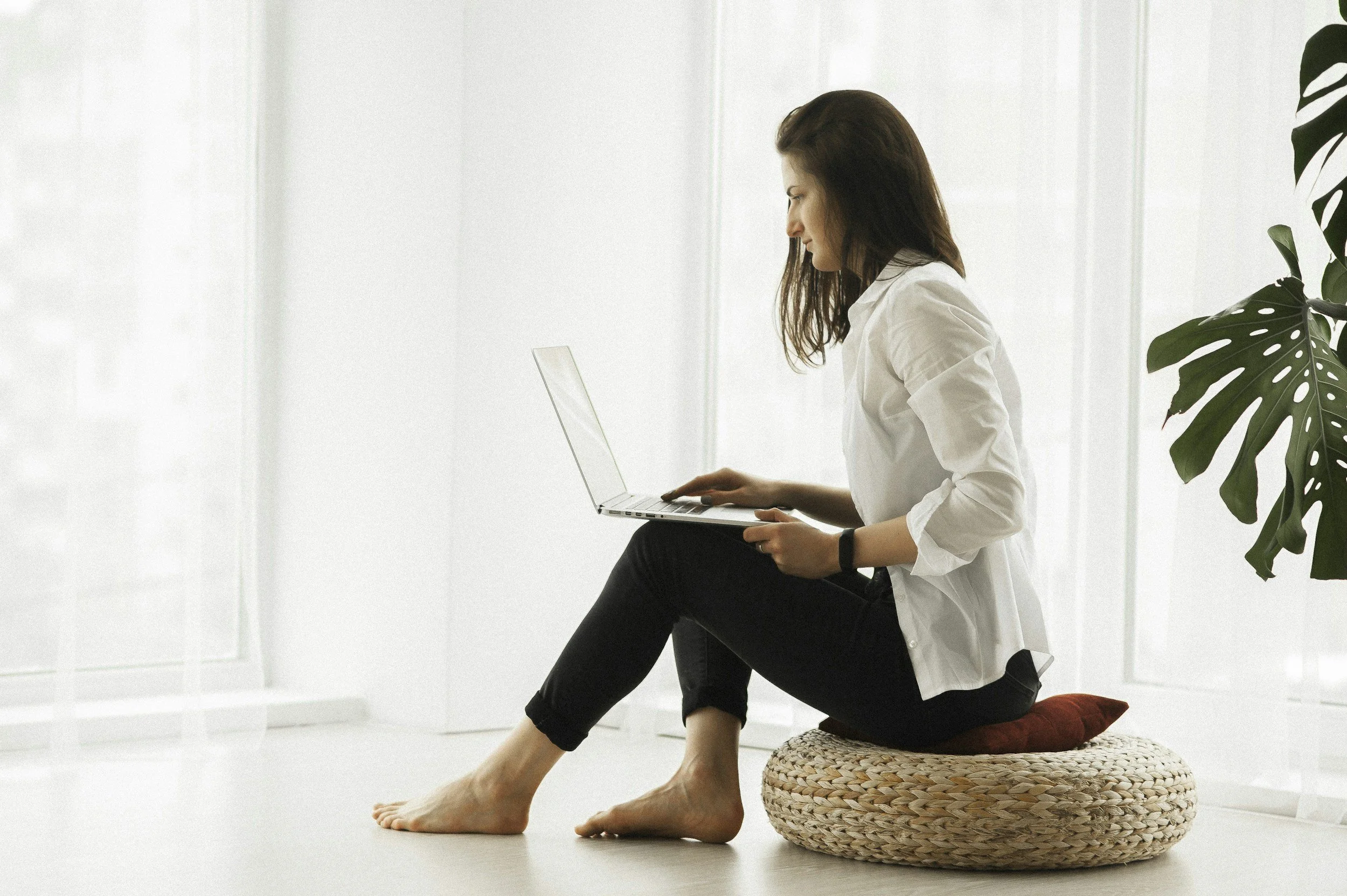 Woman sitting on a cushion on the floor using a laptop, with a large green plant nearby in a bright room.