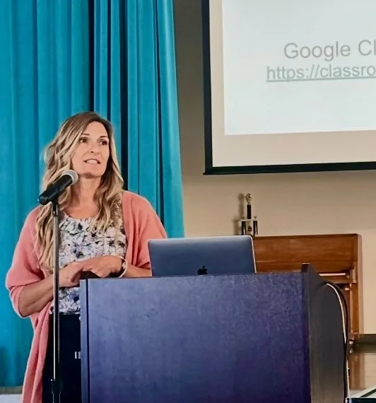 A certified somatic practitioner  stands at a podium with a laptop and microphone, giving a presentation in a room with blue curtains and a projected screen.