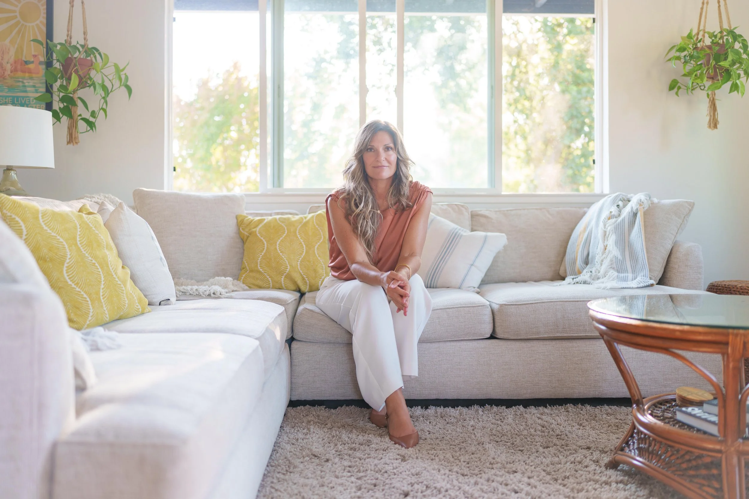 A certified somatic practitioner sitting on a beige couch with yellow and white pillows in a bright living room with large windows and green trees outside.