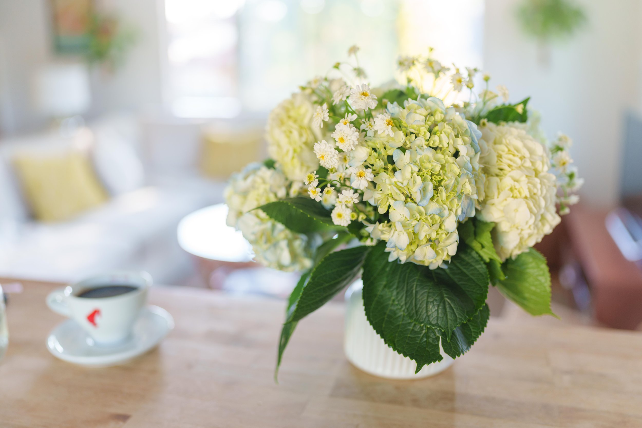 A bouquet of white and green flowers in a white vase on a wooden table, with a cup of coffee and blurred background.