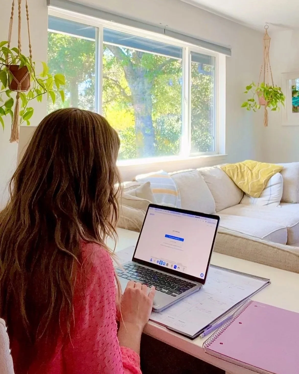 A woman with long wavy brown hair working on a laptop at a desk in a bright living room with large window, a white sofa with yellow and light-colored pillows, and hanging green plants.