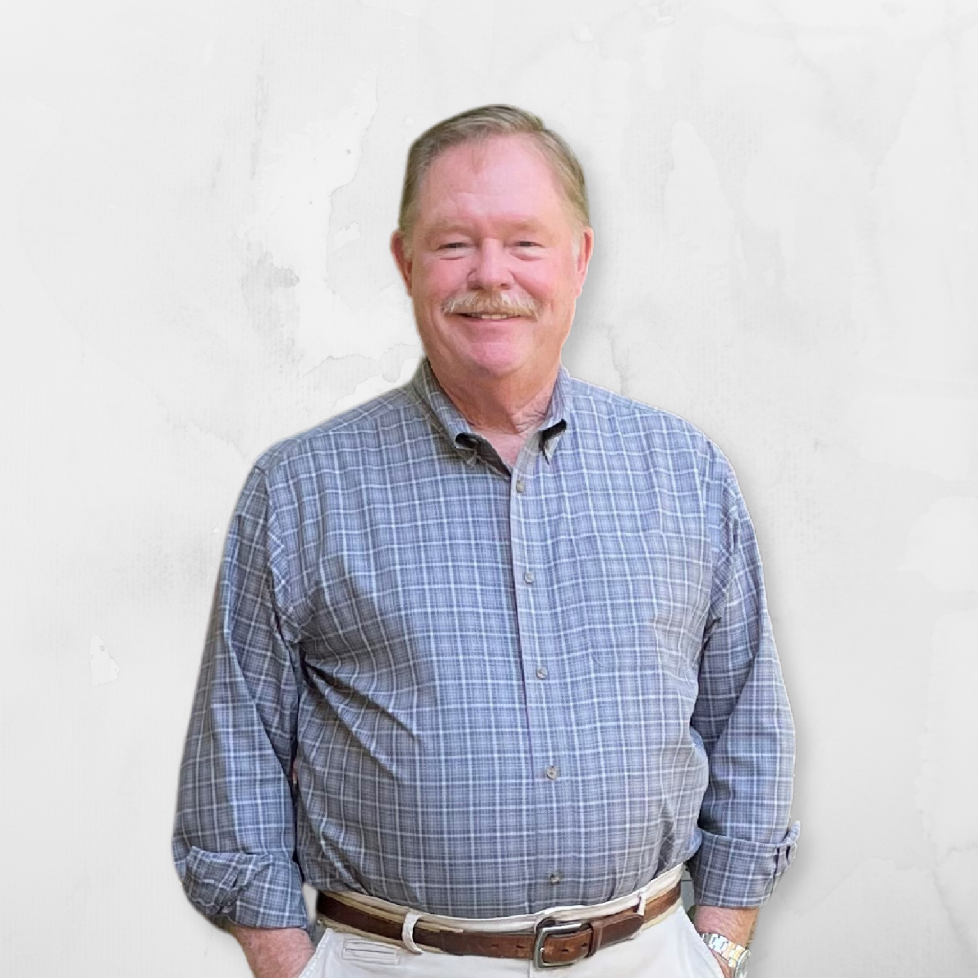 A smiling man with a mustache and short blonde hair, wearing a blue plaid shirt, beige pants, and a brown belt, standing against a plain white wall.