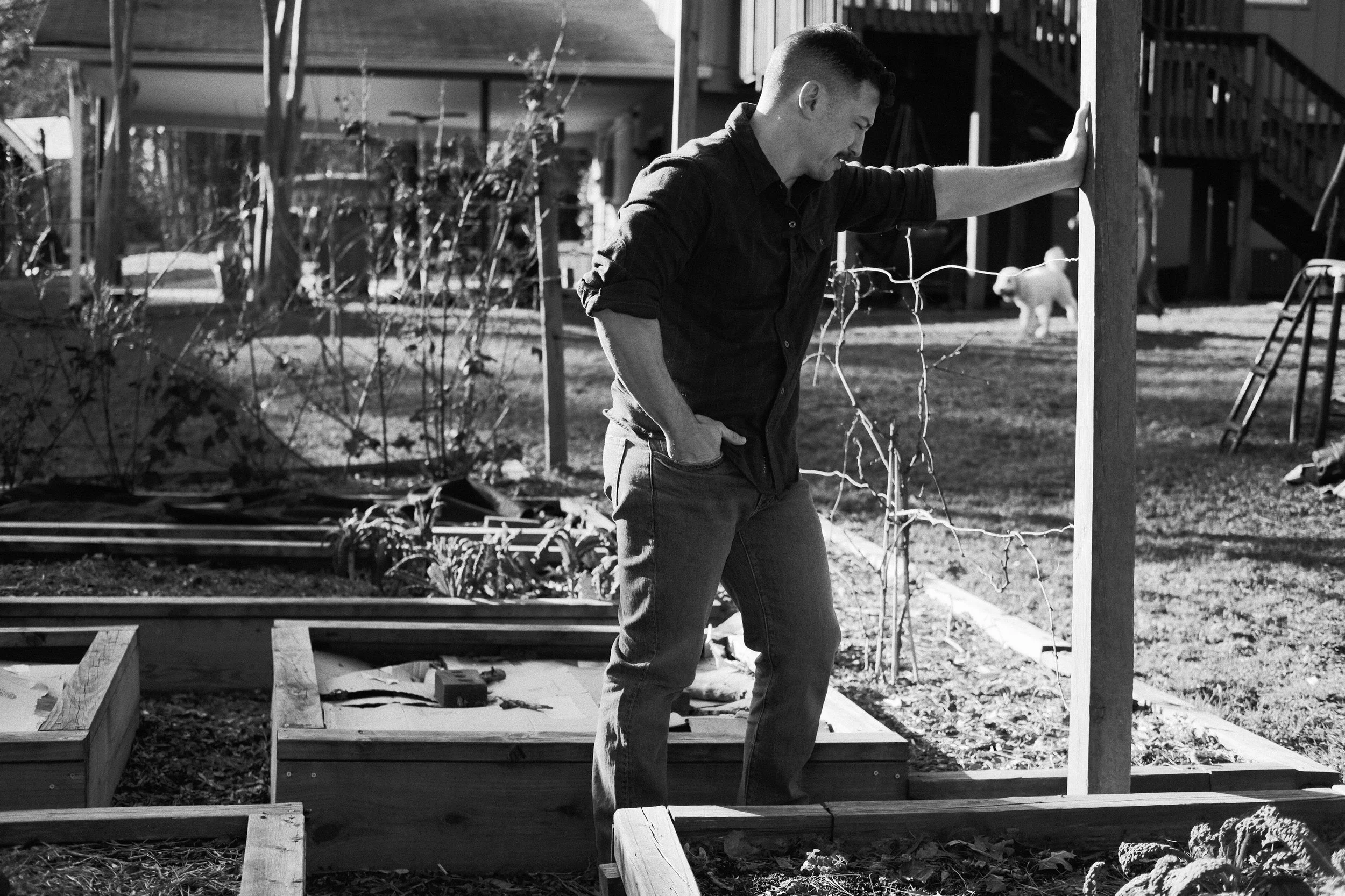 A man in a plaid shirt stands outdoors on a wooden deck, smiling with one hand on his hip and the other against a wooden post, in a residential backyard with trees, a white dog, and a staircase in the background.