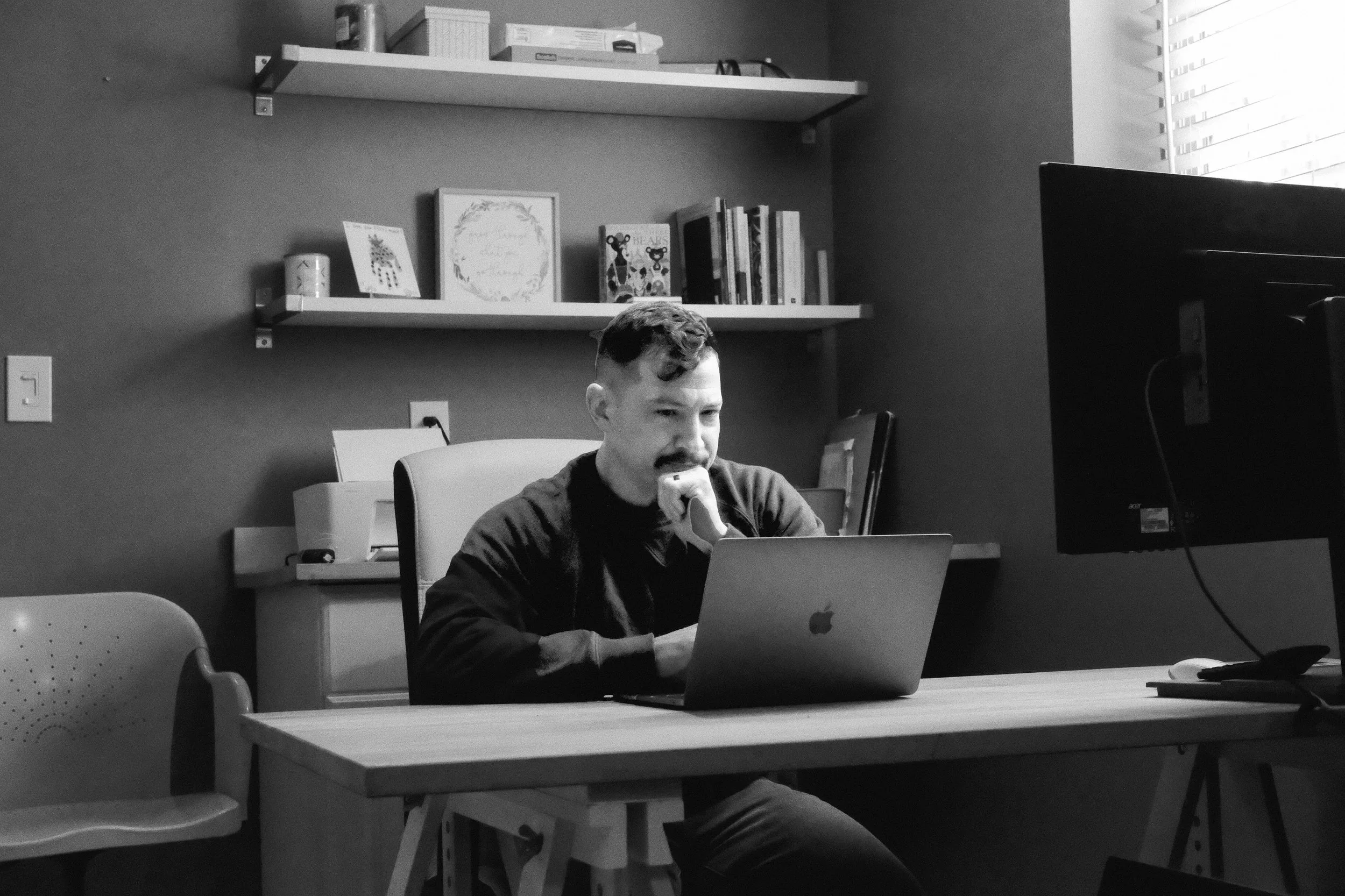 A man with a mustache sitting at a desk working on a laptop in an office. There are shelves with books and decorative items on the wall behind him, and a large window with blinds to the right.