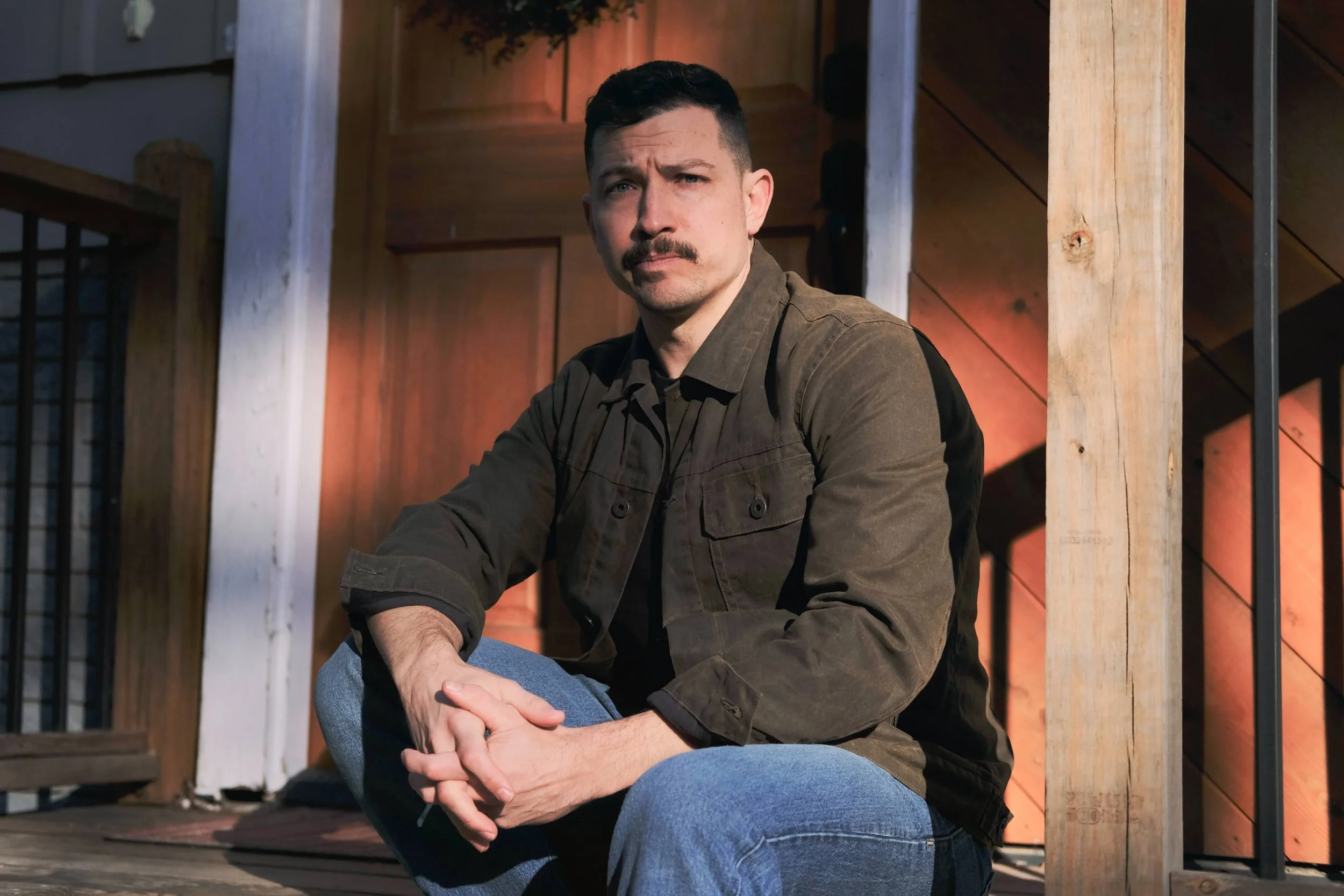 A man with dark hair and a mustache sitting outdoors on a wooden porch, wearing a brown shirt and blue jeans, looking at the camera with a serious expression.
