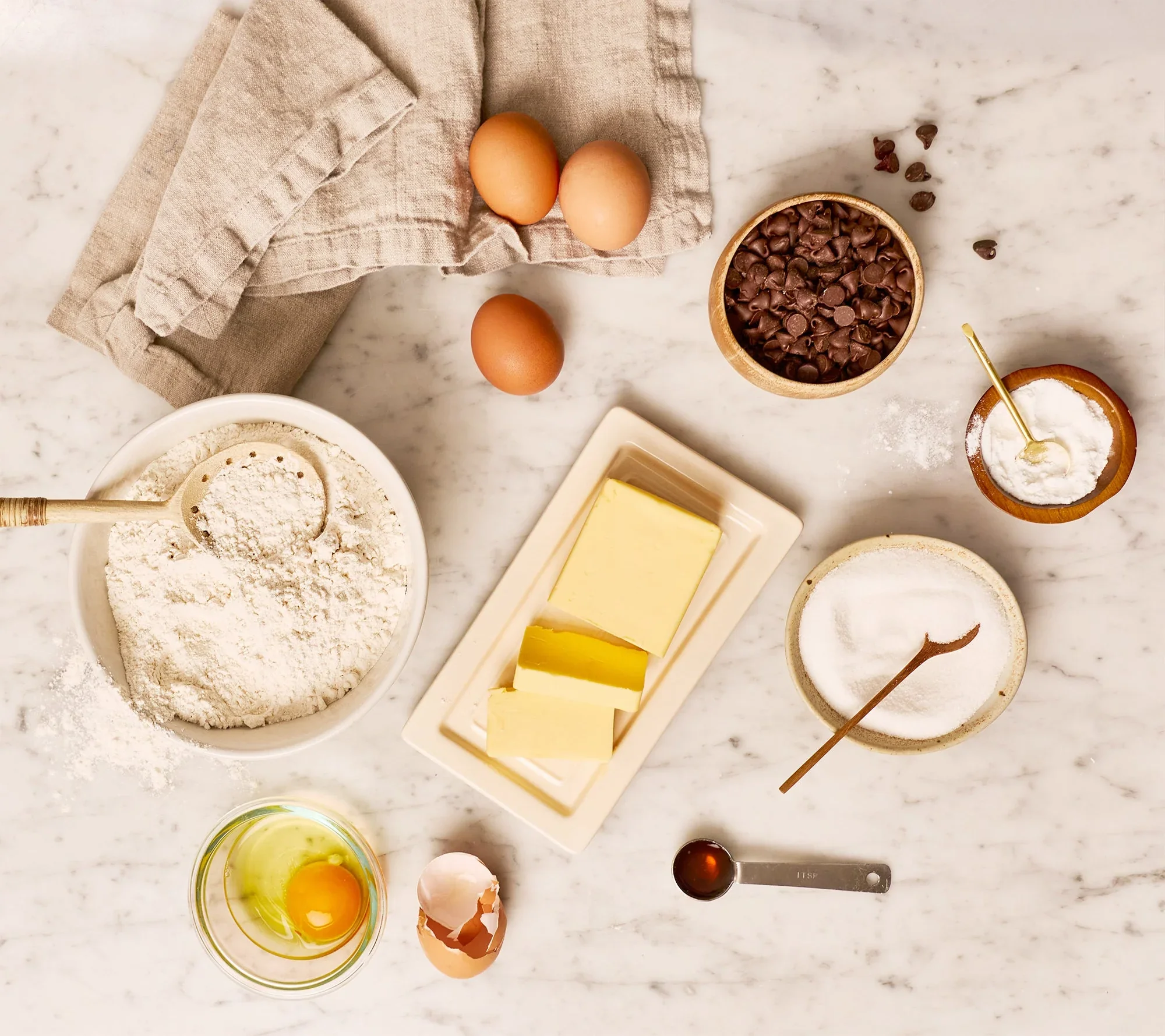 Baking ingredients on a marble countertop, including eggs, chocolate chips, flour, butter, sugar, and eggs in a bowl.