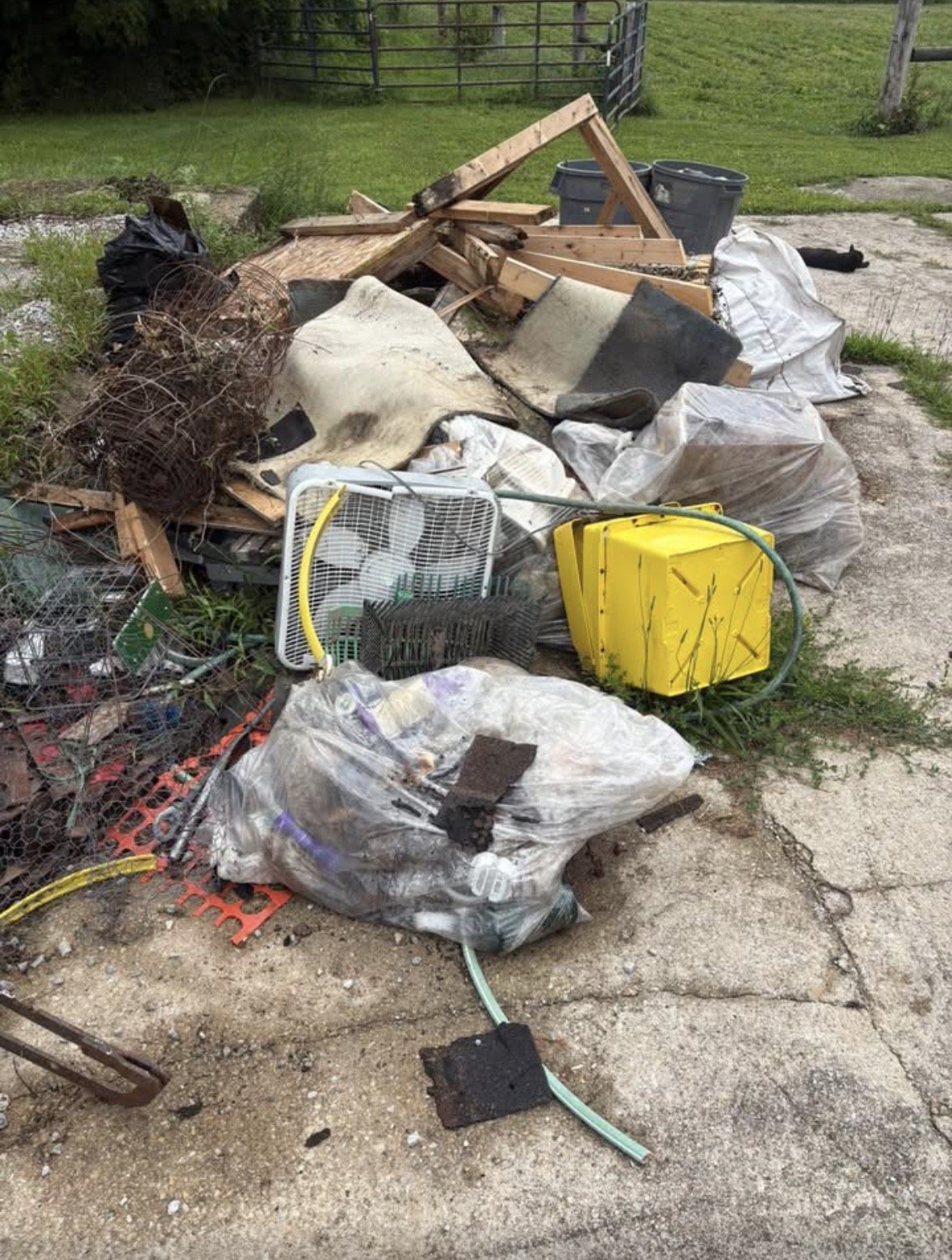 Pile of discarded trash including wood debris, plastic bags, a yellow hose reel, a small fan, and various scraps on a cracked concrete surface outdoors near a grassy area with a fence.