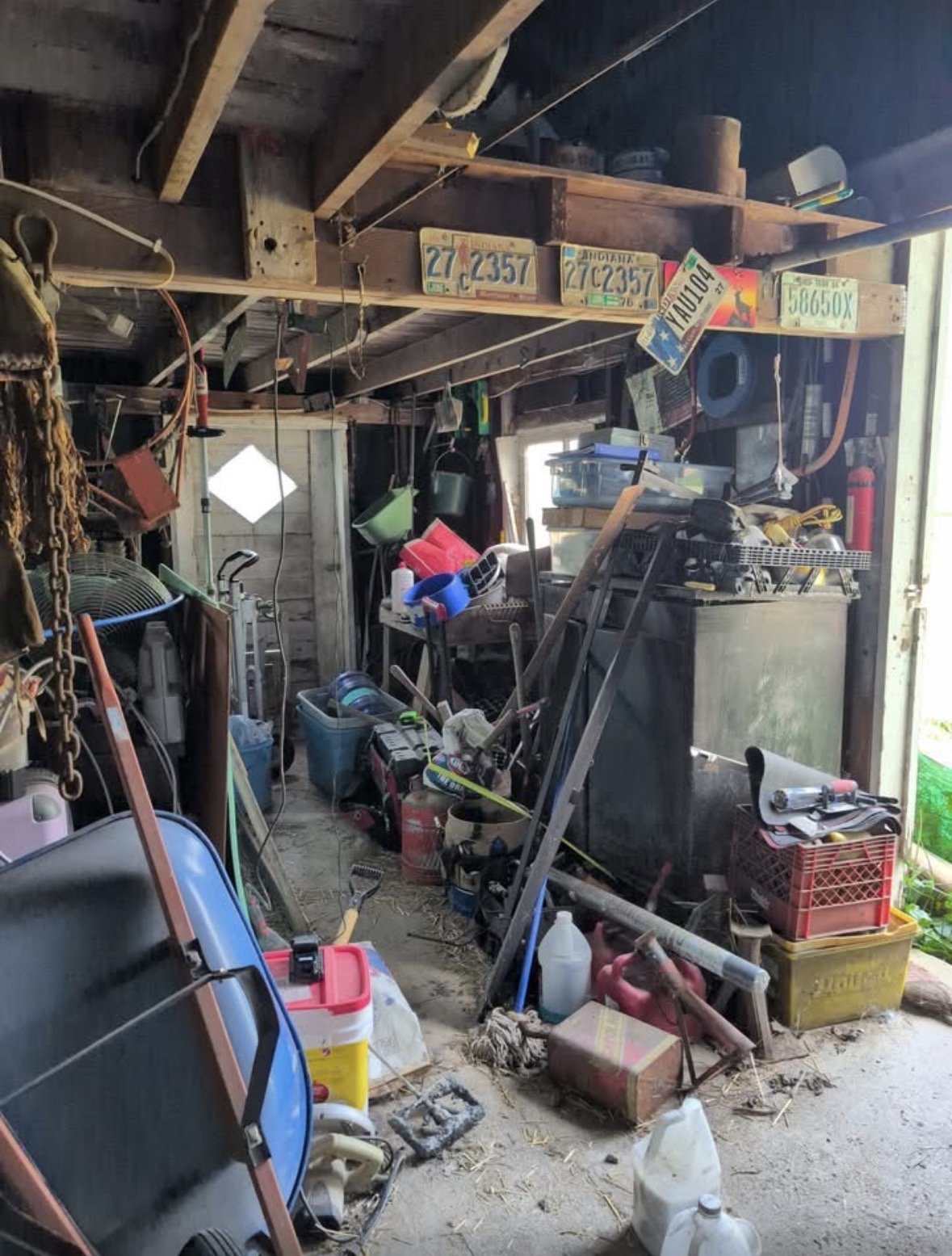 A cluttered garage filled with tools, storage bins, and miscellaneous items, with unfinished walls and a partially open door.