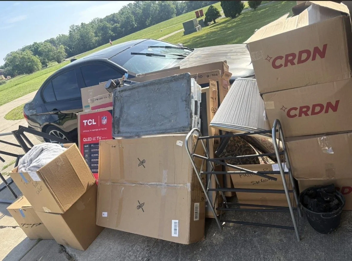 Piles of cardboard boxes, a shopping cart, and various household items placed on a sidewalk next to a parked black car, with grassy park area and trees in the background.