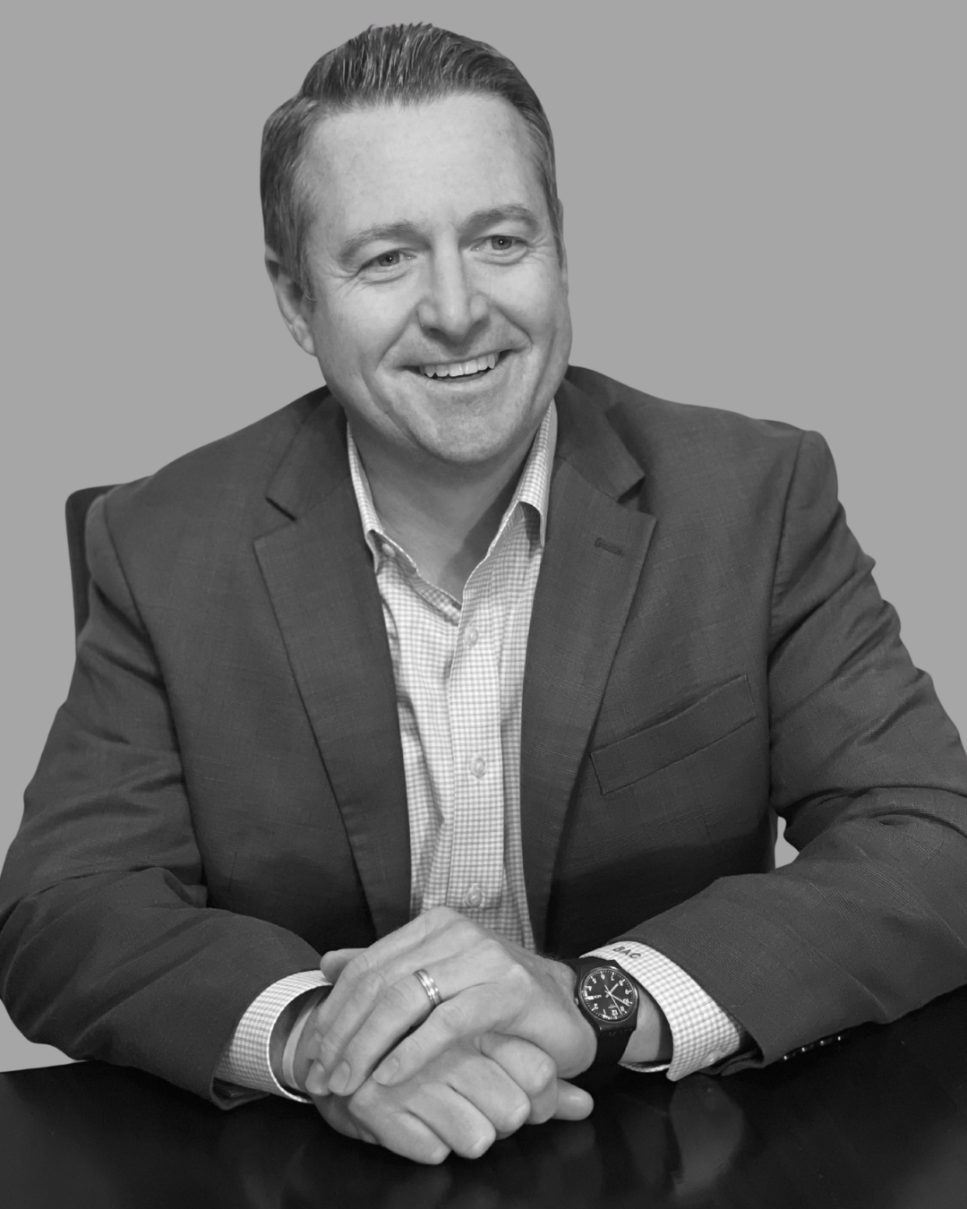 A black and white photo of a smiling man in a suit, sitting at a table with his hands clasped, wearing a watch and a wedding band.