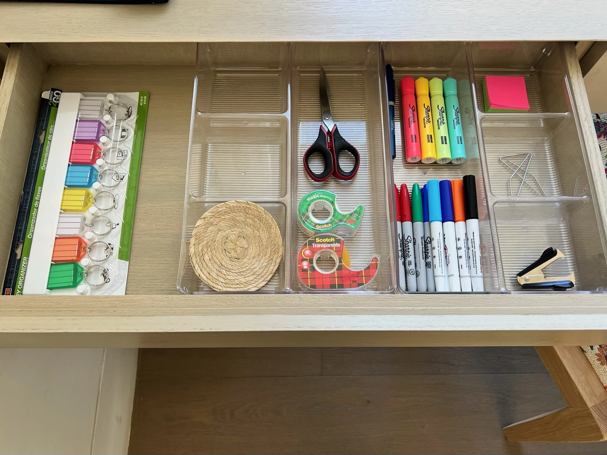 A partially opened desk drawer containing office supplies, including highlighters, markers, sticky notes, scissors, tape, and paper clips.