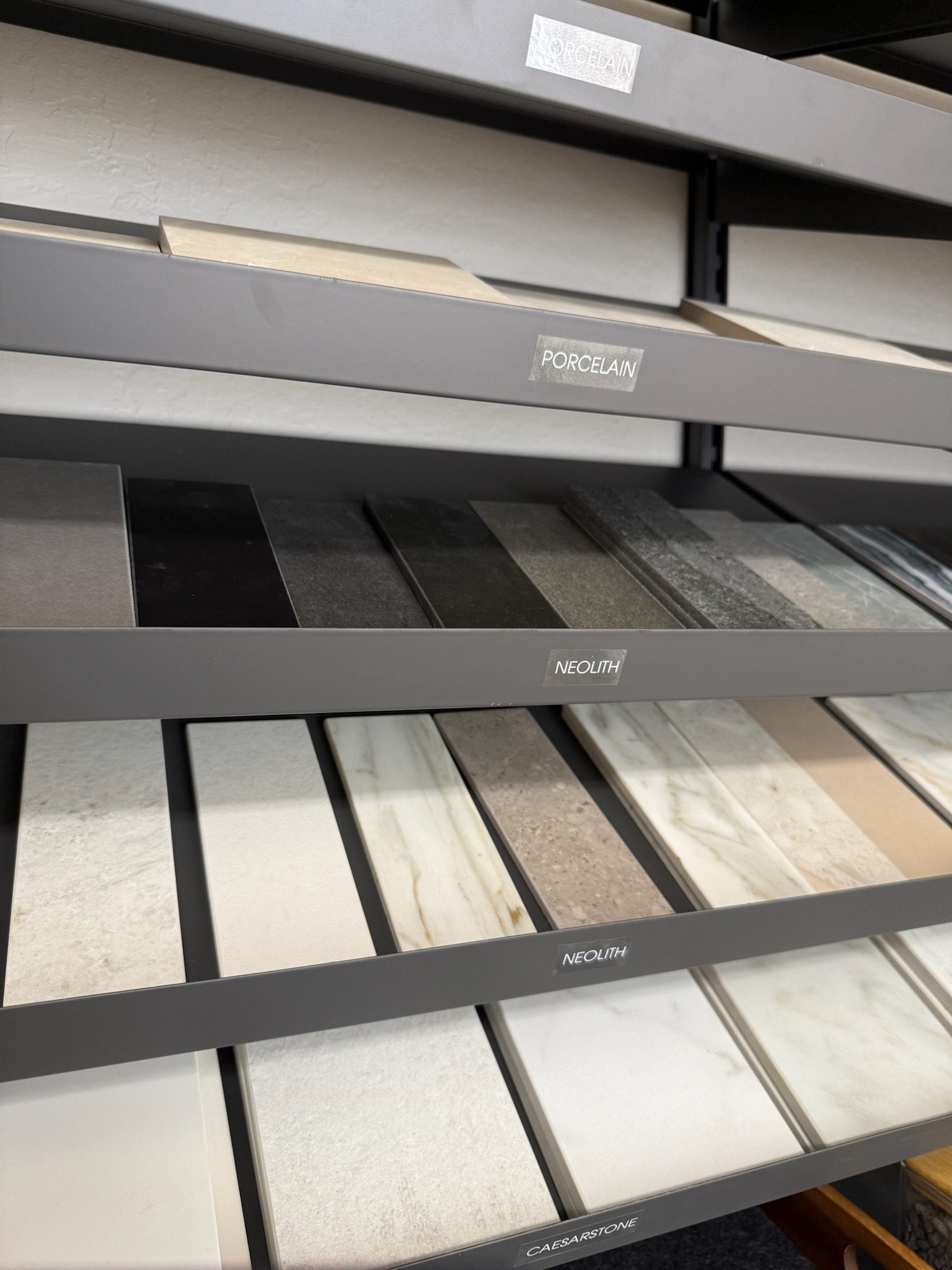 Display of various marble and porcelain tile samples on a retail shelf, labeled with brands 'Porcelan,' 'Neolith,' and 'Caesarstone'.