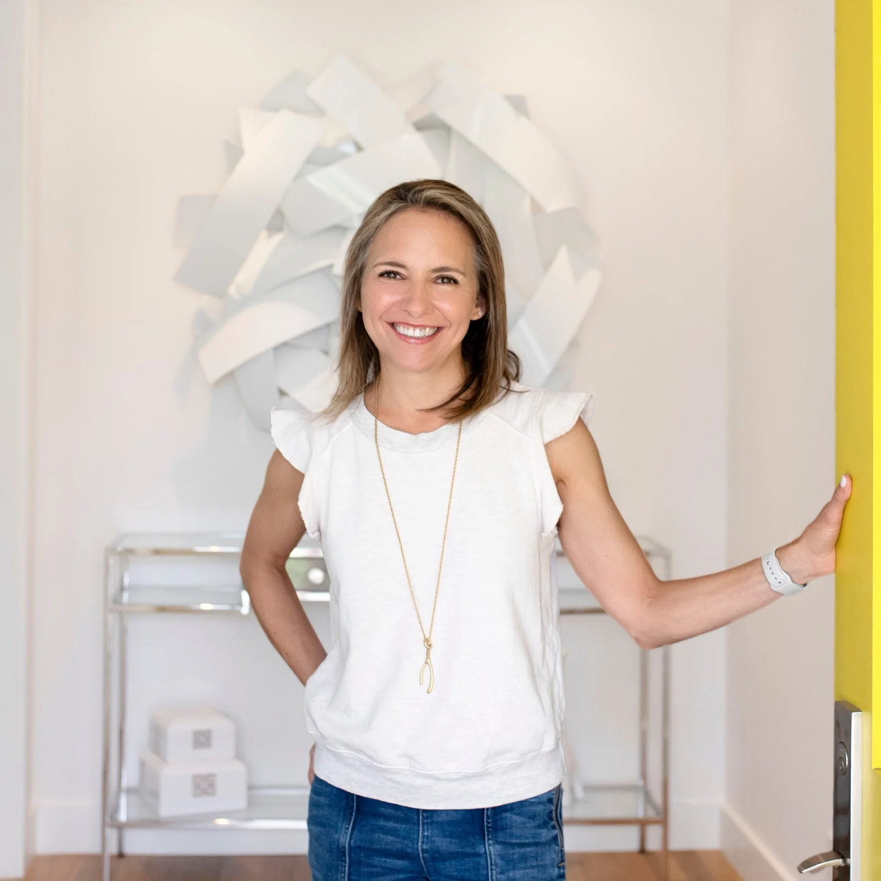 Smiling woman in white top and jeans standing near yellow door, holding the door handle with her right hand, in a bright room with white walls and a shelving unit in the background.