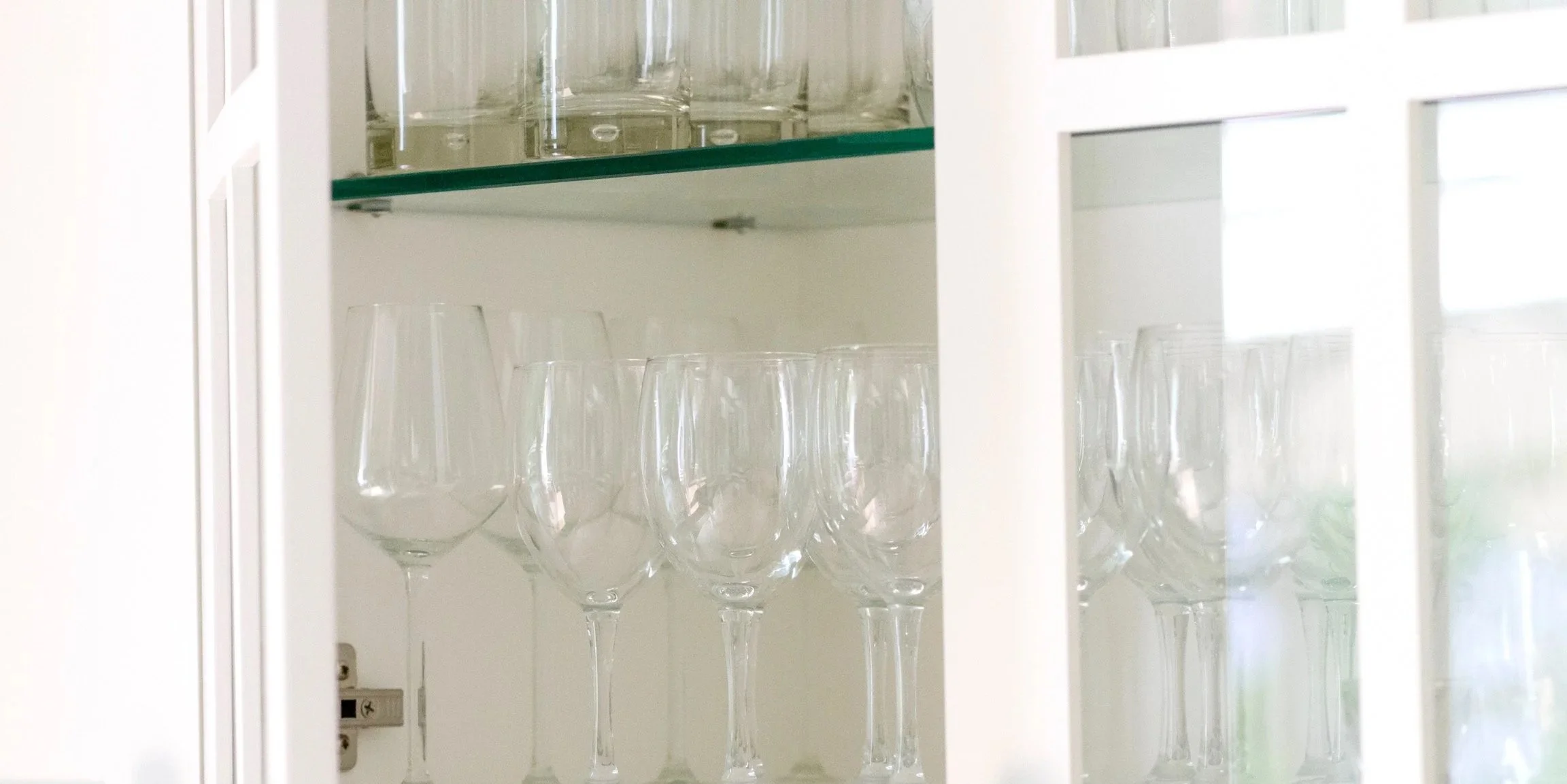 Inside a white cabinet with two shelves, the top shelf holds empty glasses and the bottom shelf has wine glasses.