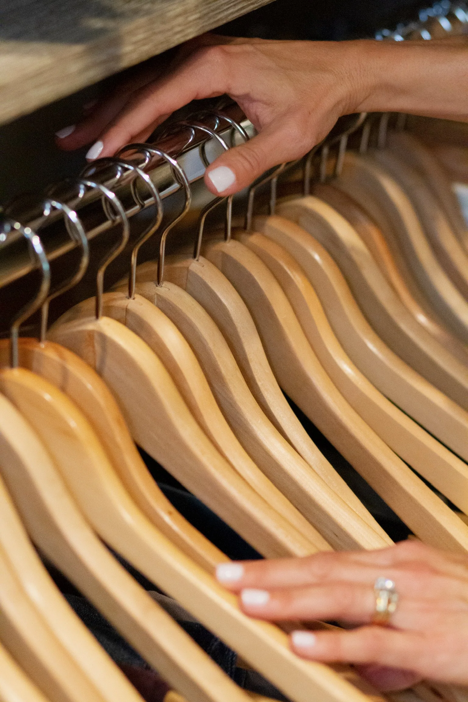 A person's hand with white nail polish reaches into a closet or wardrobe to grab a wooden hanger from a row of similar hangers.