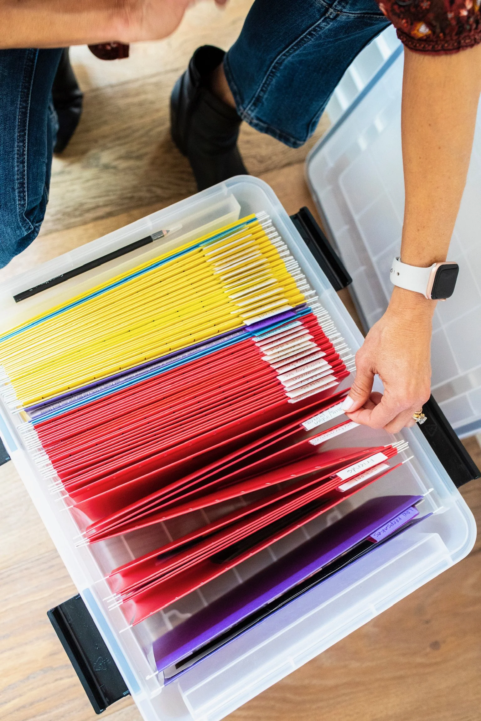 A person is organizing colorful files in a plastic storage container. The files are in yellow, red, purple, and black colors, with some labeled tabs. The person's arm is visible, wearing a white wristband and a decorative ring, and they are wearing casual clothing.