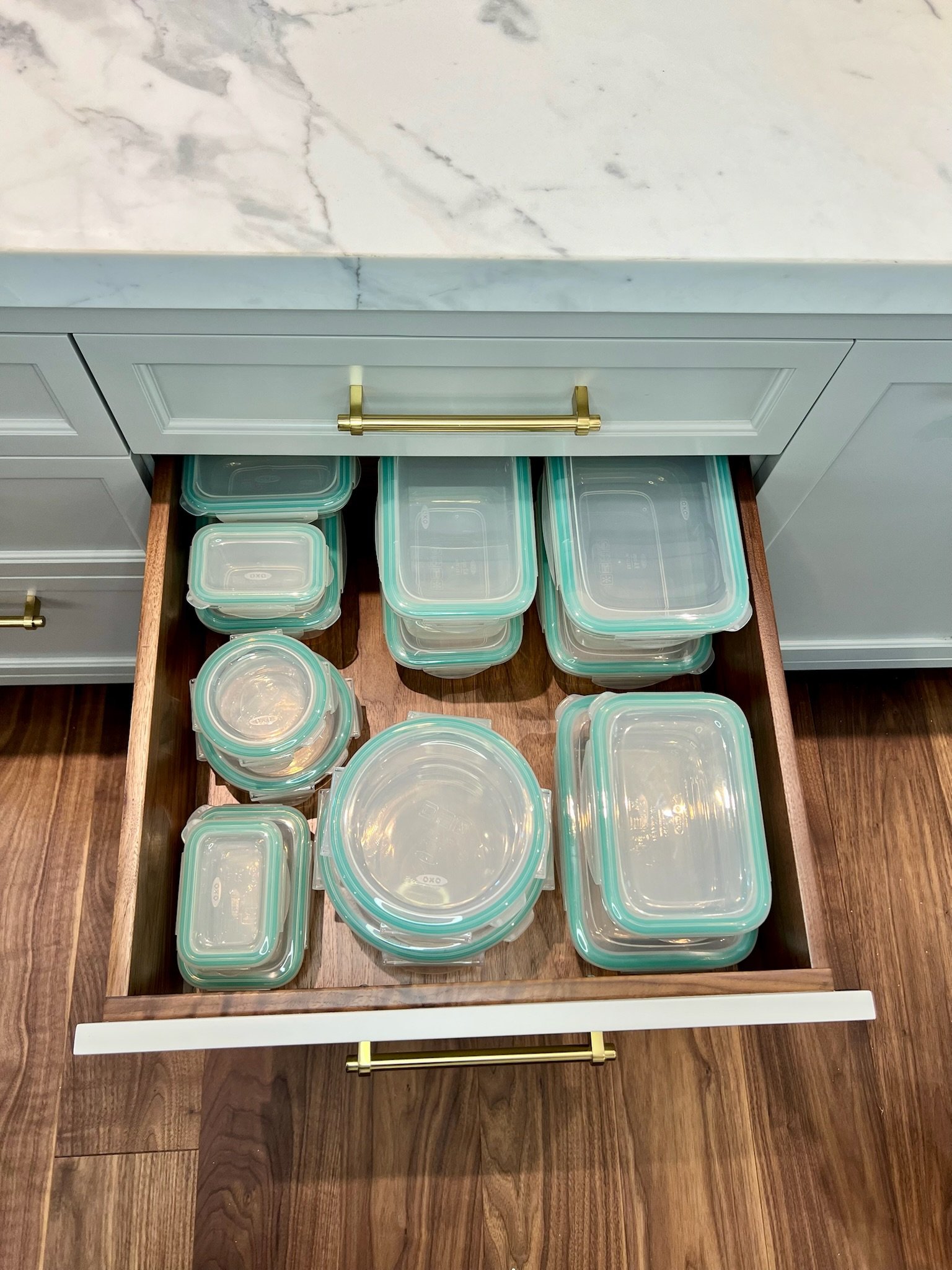 An open kitchen drawer filled with stacked glass food storage containers with teal lids, seen from above on a wooden floor.
