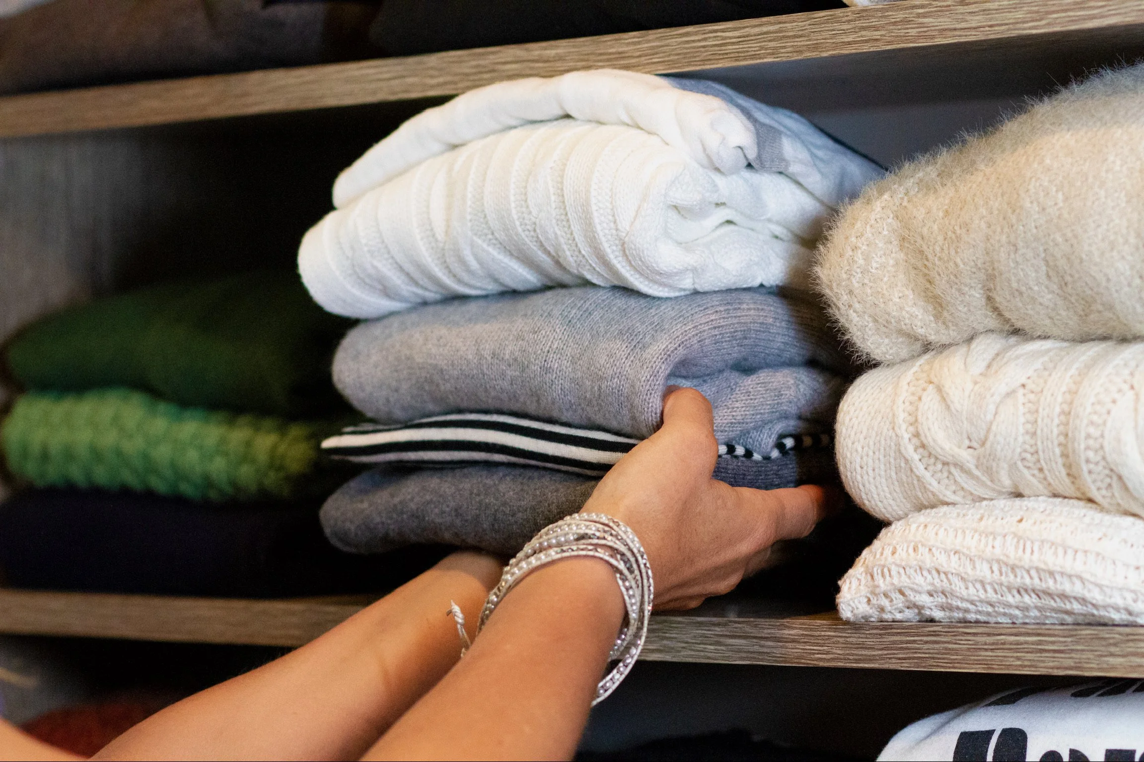 Person's hand touching neatly folded sweaters on a dark wooden shelf.
