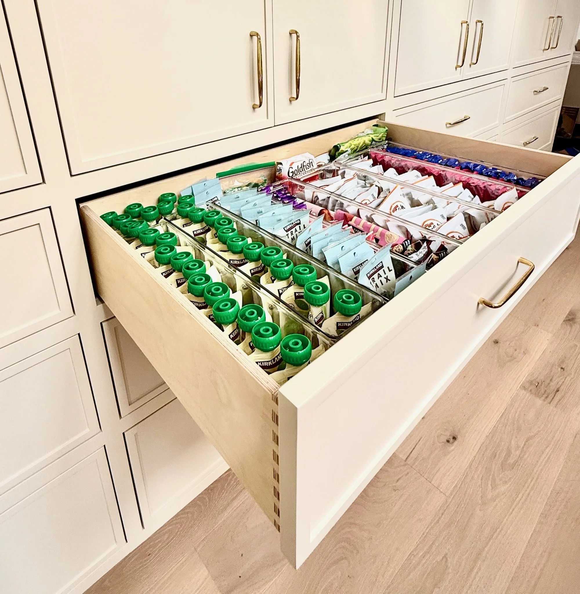 Drawers filled with various snacks, including green bottles, snack packages, and other items, in a kitchen cabinet.