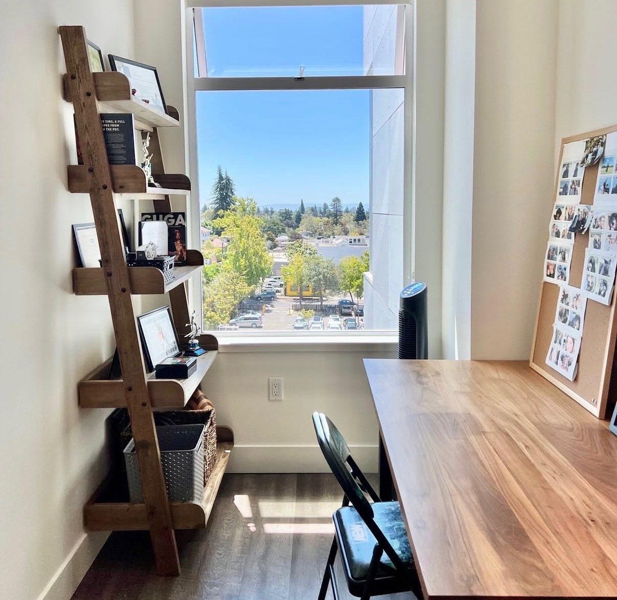 A bright office space with a large window showing a parking lot and trees outside, a wooden desk with chairs, a corkboard with photos, and a wooden shelving unit with pictures and decorative items.