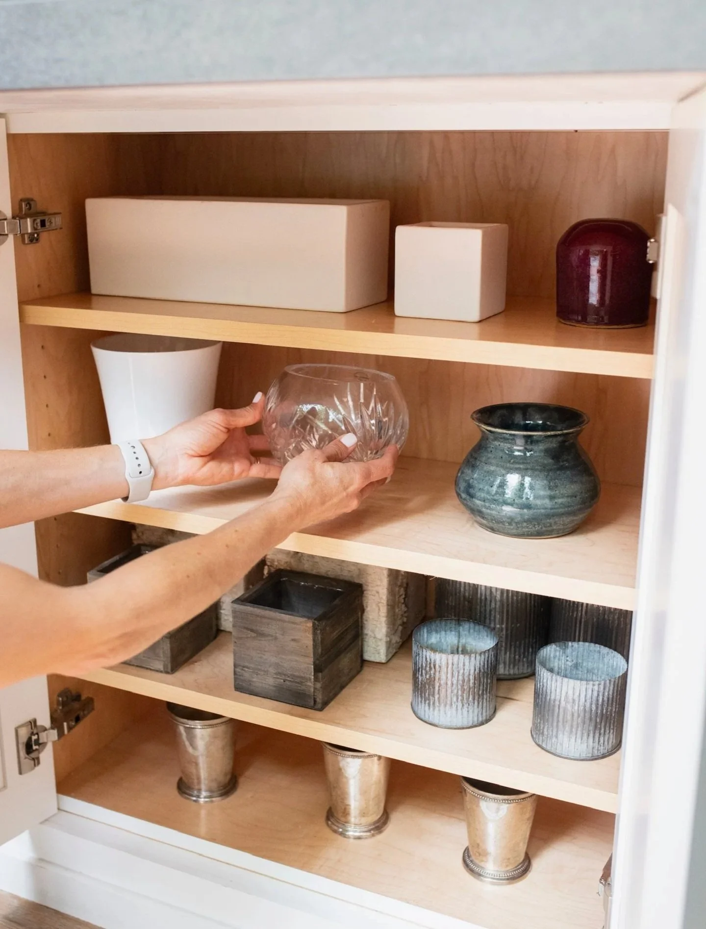 A person organizing a glass bowl in a wooden cabinet with shelves containing various decorative vases and containers.