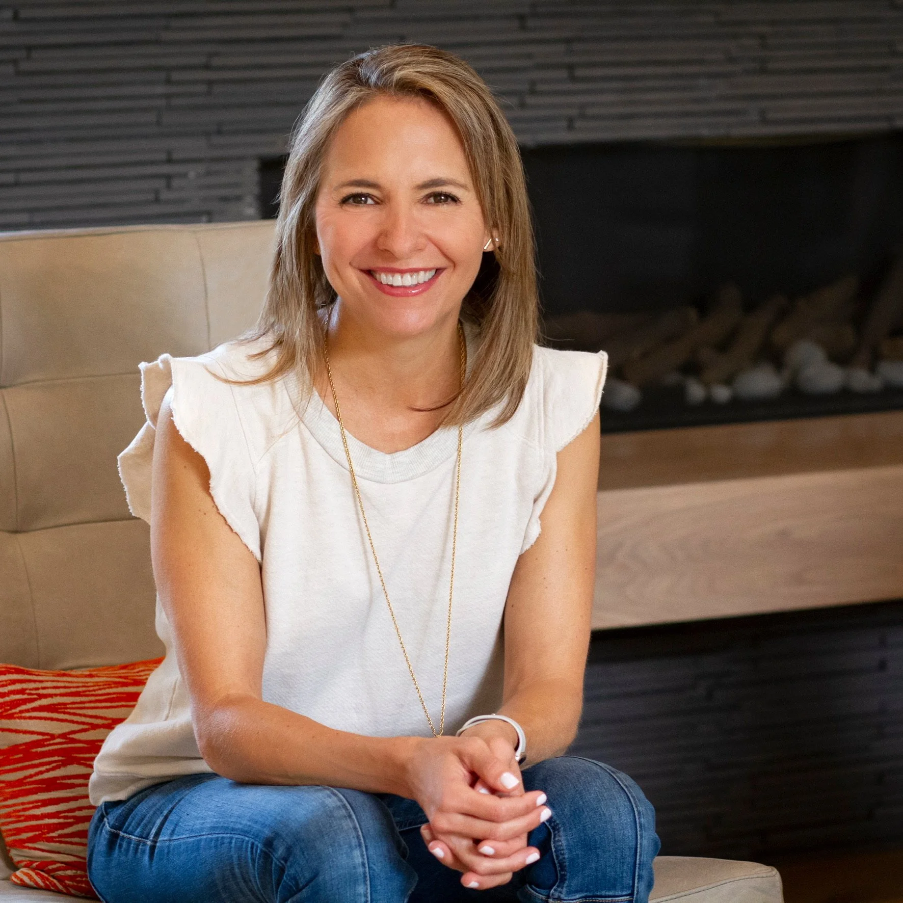 Smiling woman sitting on a beige chair in front of a modern fireplace with black horizontal tiles, wearing a sleeveless white top and blue jeans.