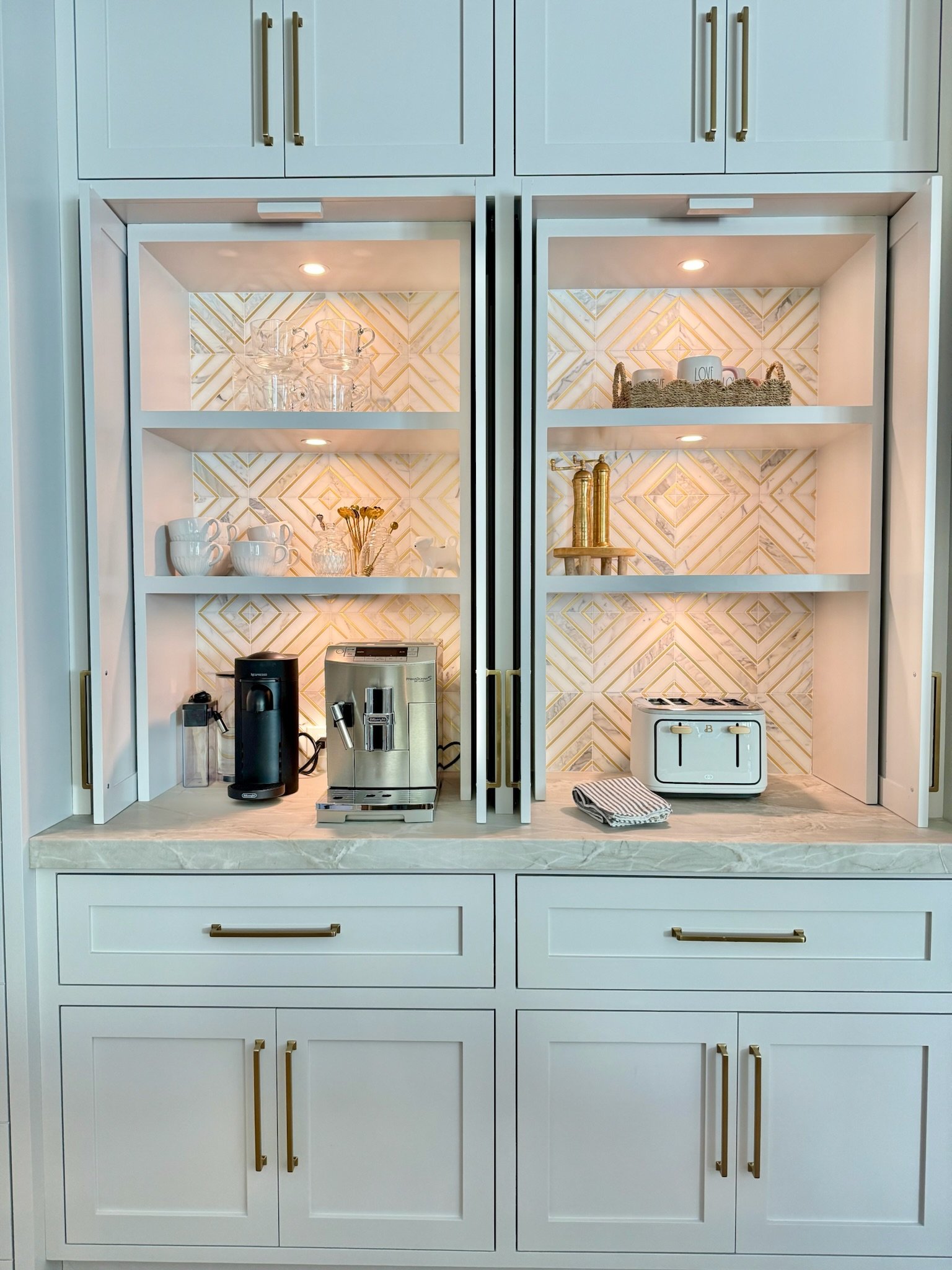 A white kitchen cabinet with gold handles, featuring open shelves illuminated by small lights. The top shelf has glass cups, the middle shelf has white mugs and dried flowers in a small vase, and the bottom shelf has a coffee maker, espresso machine, gold decorative items, a small toaster, and a striped towel.