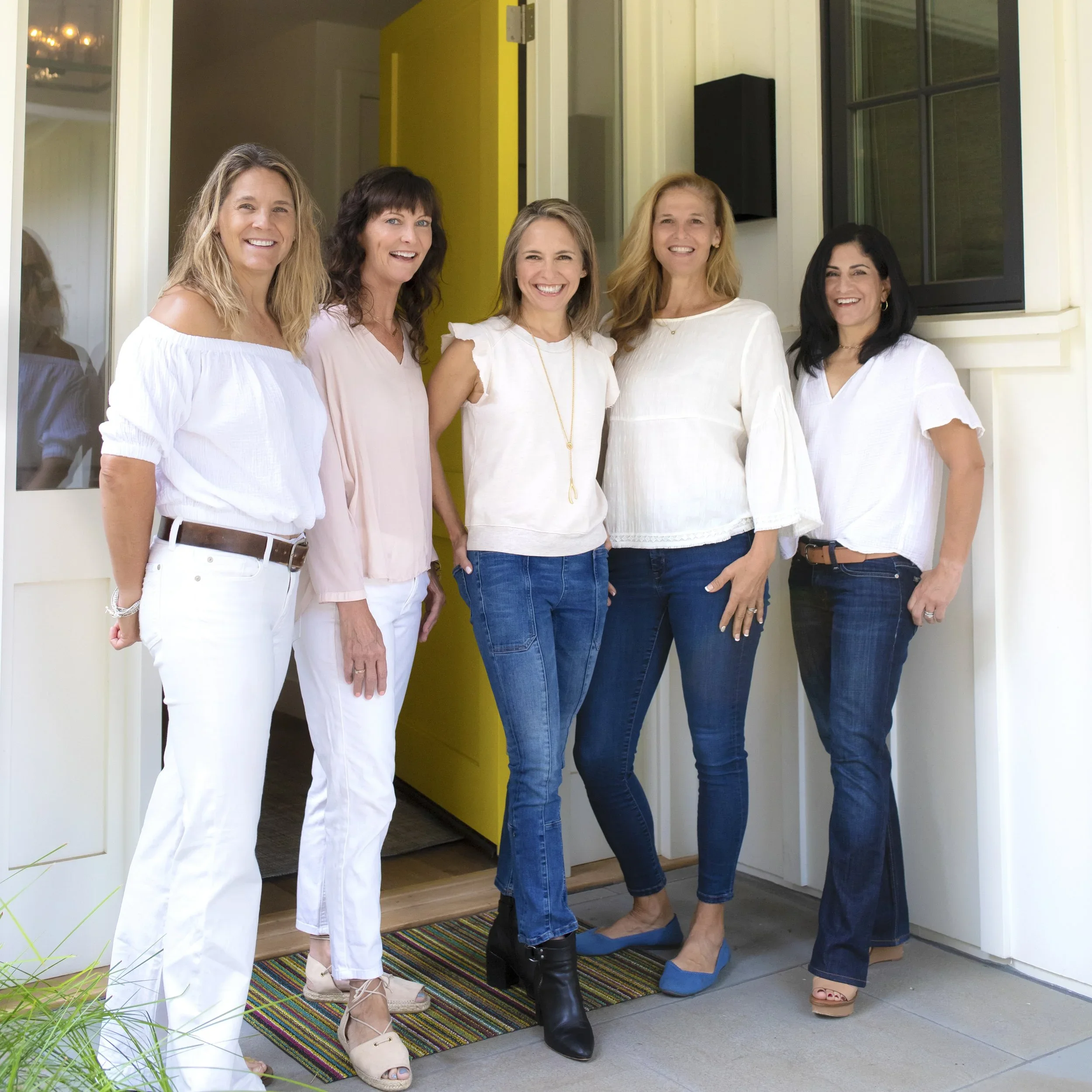 Five women standing on a porch, smiling and posing for a photo, wearing white and light-colored tops, with some wearing boots and high heels.