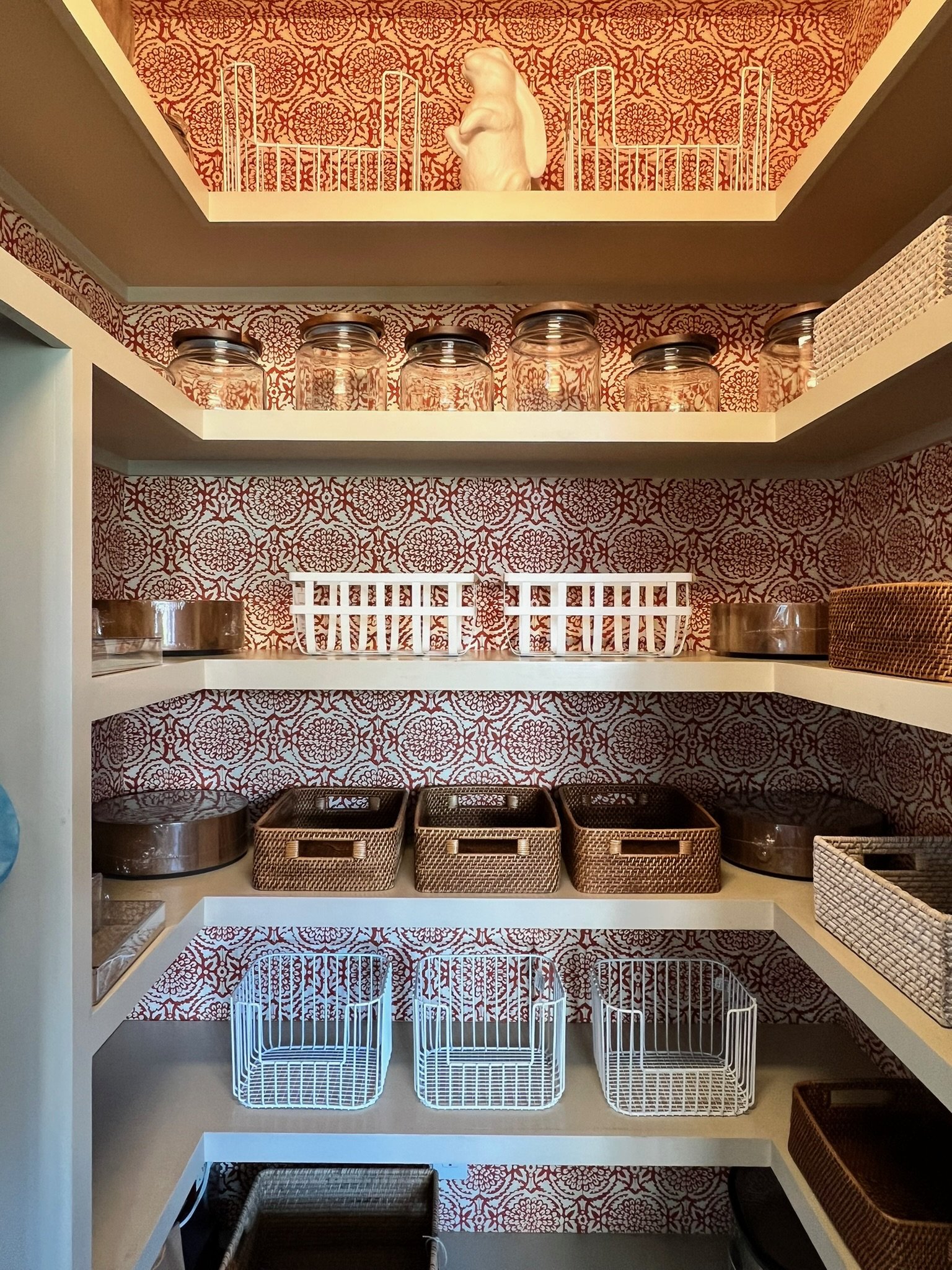 A kitchen cabinet with three shelves filled with storage containers and baskets, decorative red and white wallpaper, and a small sculpture of a seated figure on the top shelf.