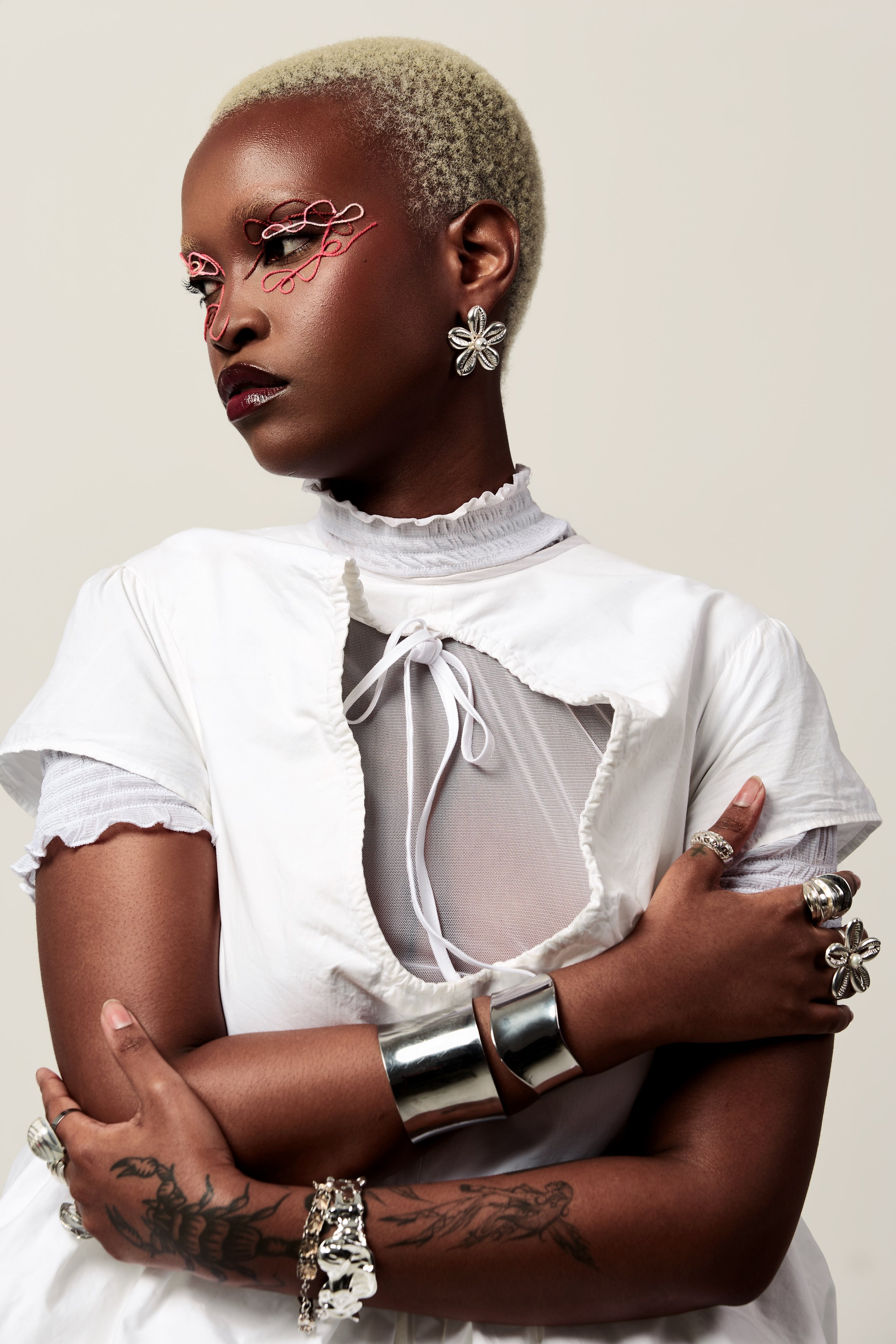 Portrait d'une femme noire avec une coupe de cheveux blonde très courte, portant du maquillage artistique autour des yeux, des boucles d'oreilles en forme de fleur, une blouse blanche avec un col à volants, et plusieurs bijoux, incluant des bracelets