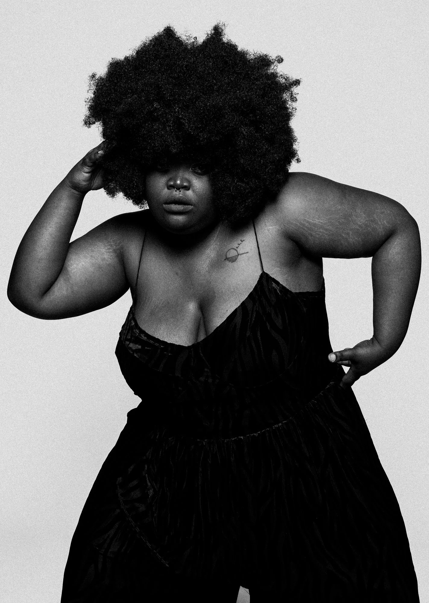 Une femme portant un haut noir avec un tatouage, avec une coiffure volumineuse et bouclée. Elle a un piercing au septum.