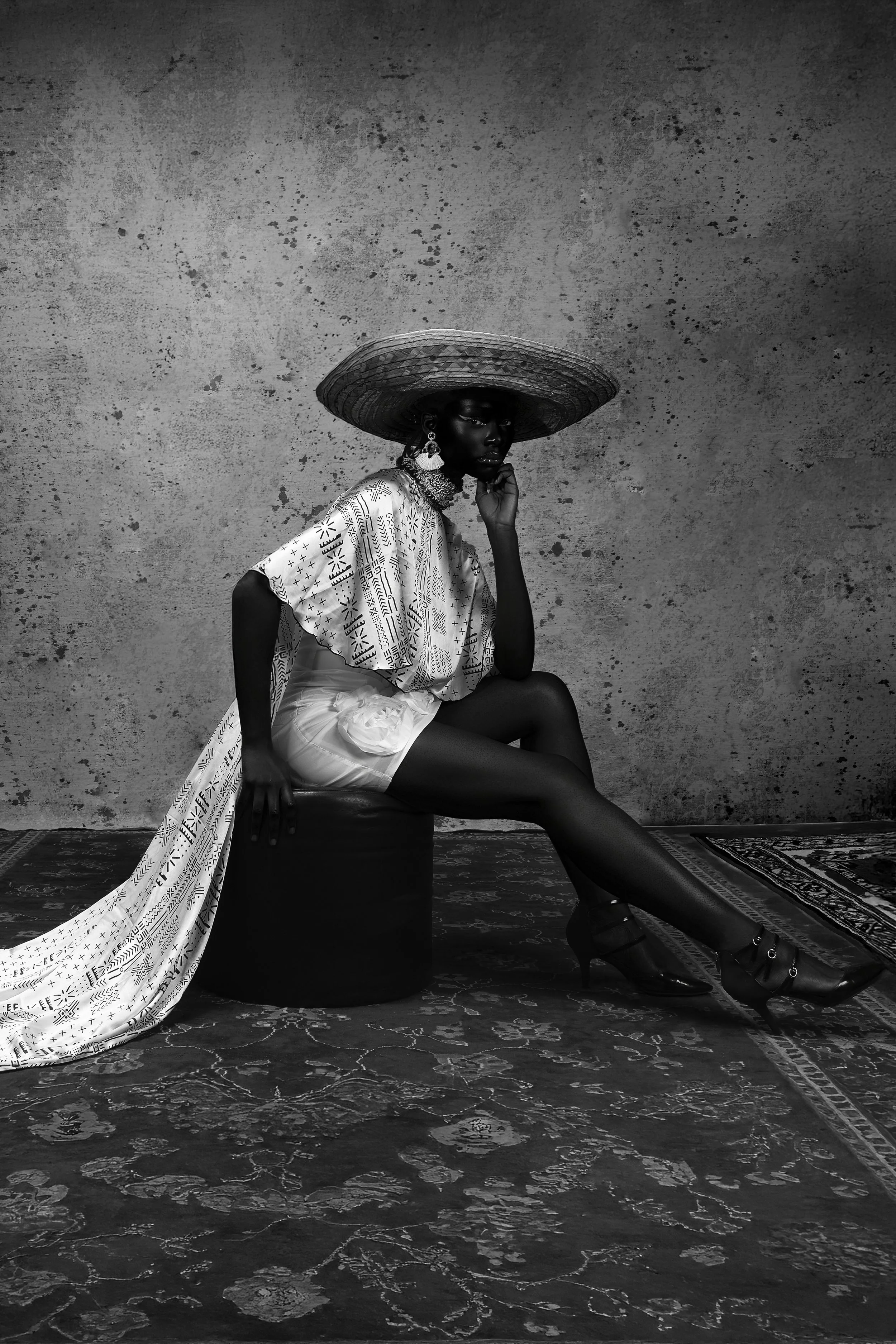 Une femme noire élégante porte un chapeau large en osier, une robe imprimée et des talons hauts, assise sur un tabouret devant un mur texturé, en noir et blanc.
