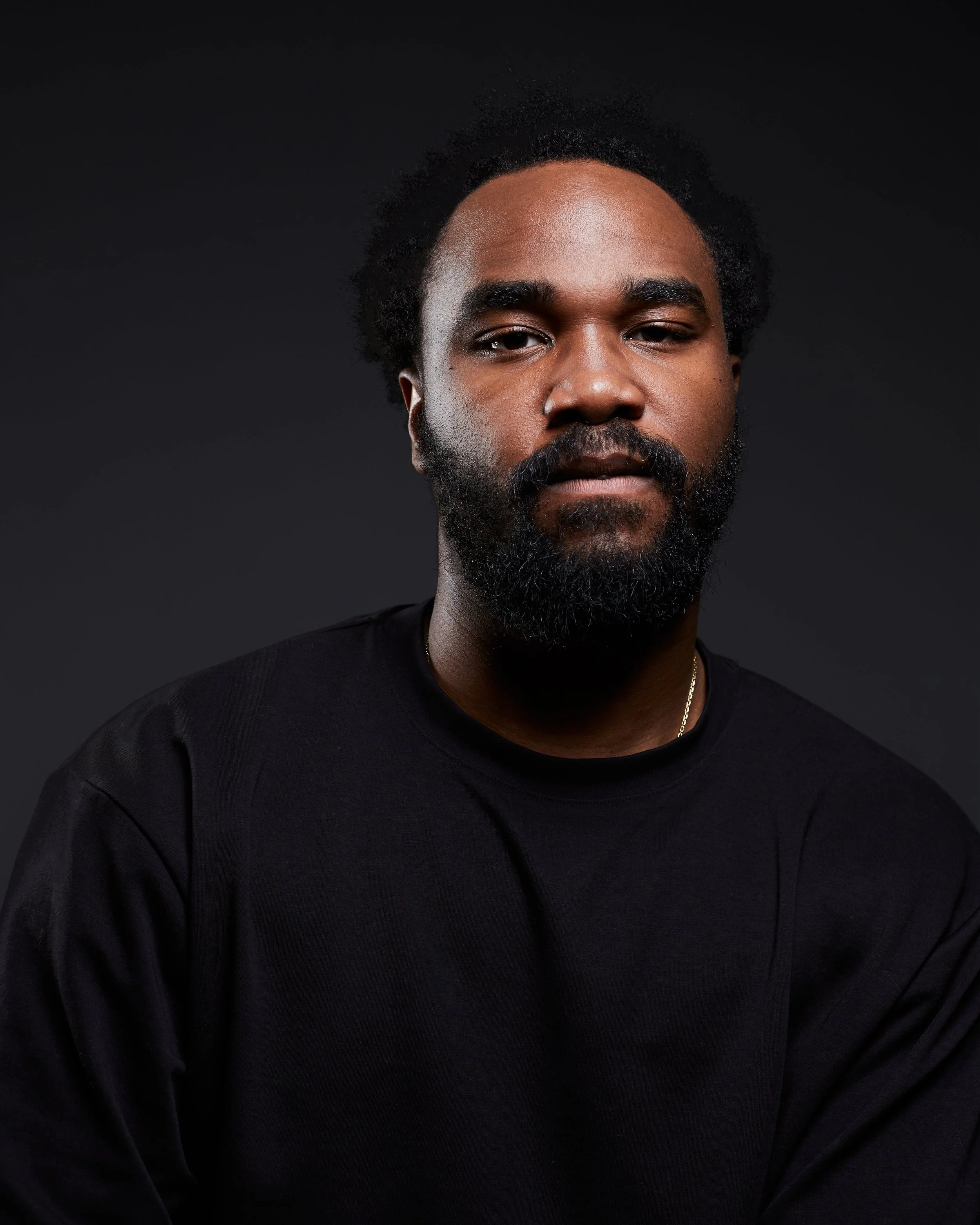 Portrait d'un homme au visage en gros plan, avec une barbe noire et des cheveux bouclés, portant un t-shirt noir, contre un fond noir.