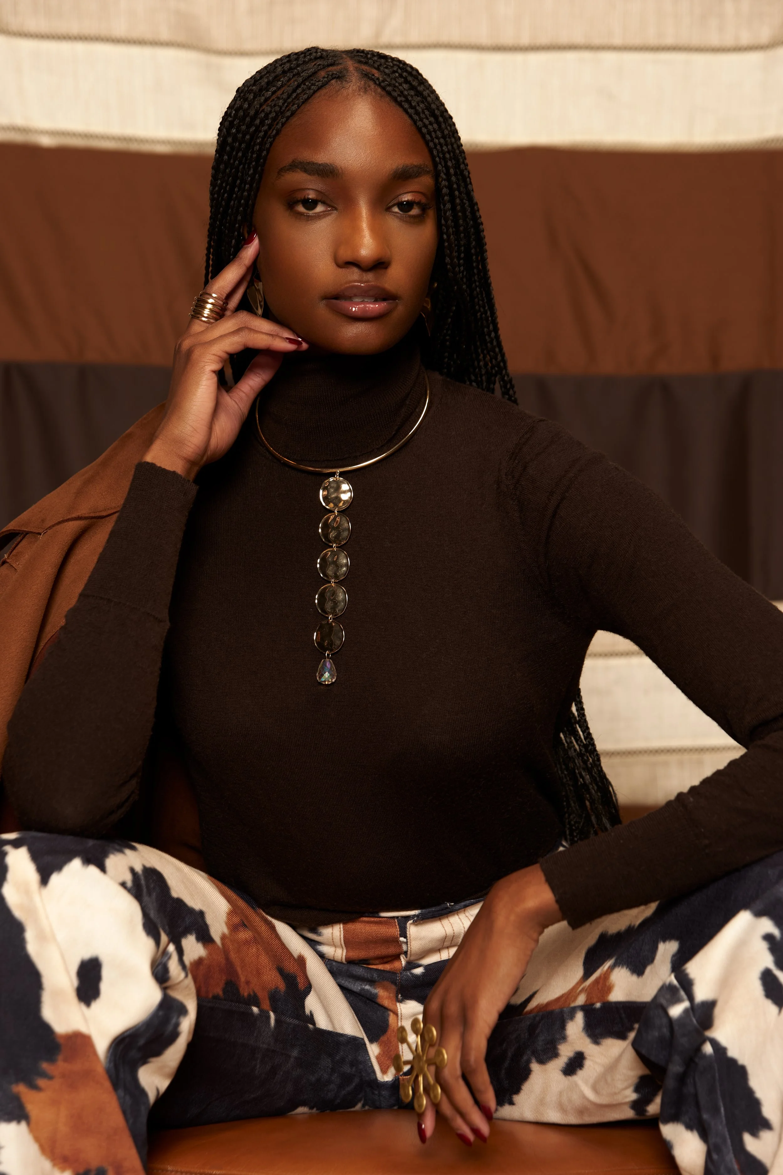 Jeune femme noire avec des cheveux en tresses porte un t-shirt noir et des bijoux, assise sur un canapé, avec un fond beige et marron.