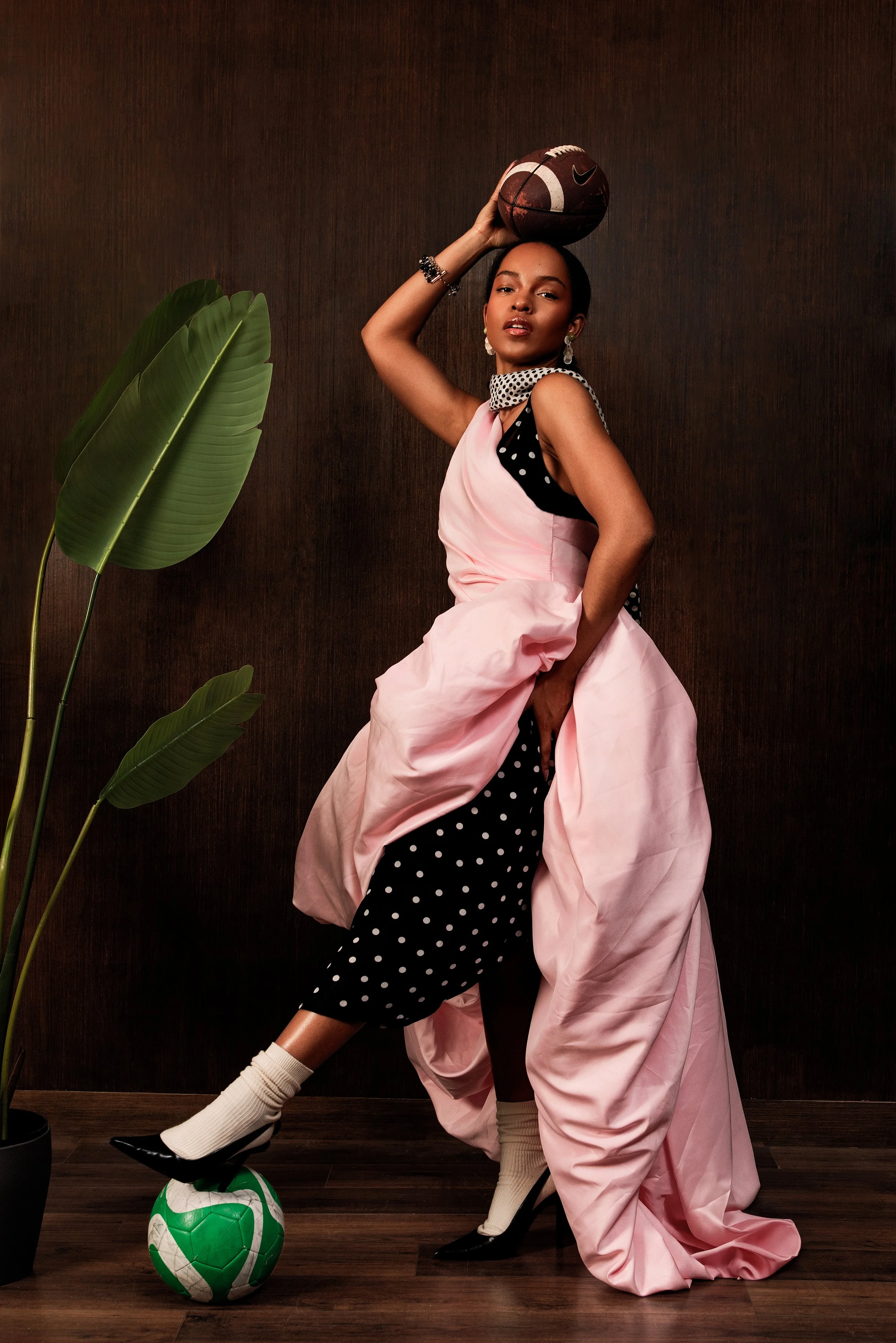 Une femme posant avec un ballon de basketball sur la tête, portant une robe rose et une autre robe noire à pois, avec des chaussures à talons noirs et des chaussettes beiges, accompagnée de plantes vertes à feuillage large dans le fond.
