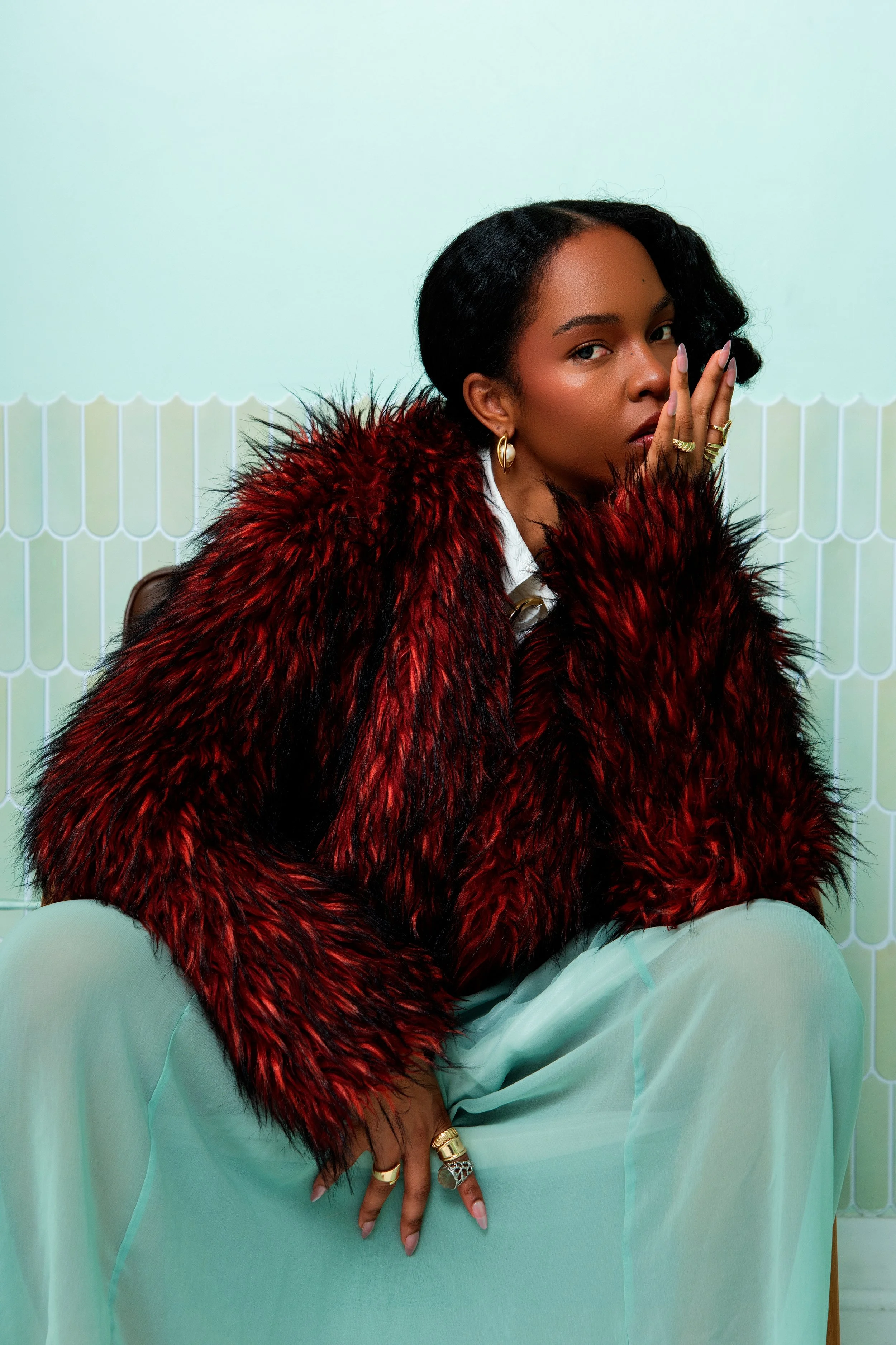 Femme portant un manteau en fausse fourrure rouge et noire avec un vêtement vert pastel, assise devant un mur à motif blanc.