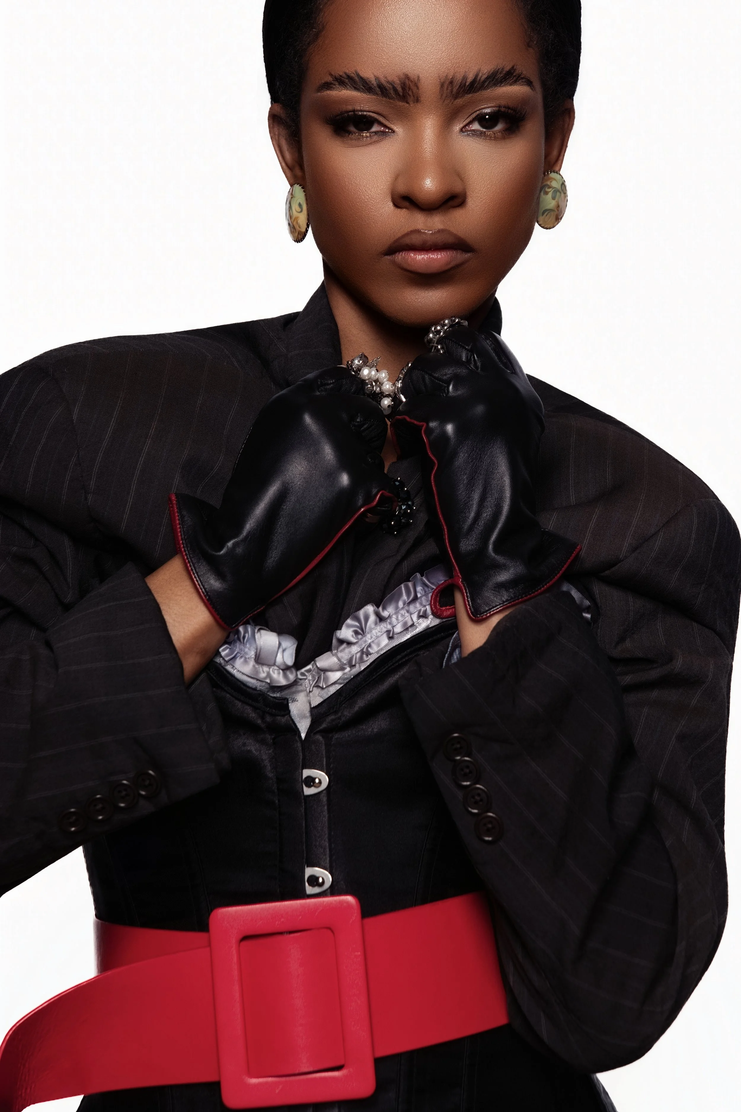 Portrait d'une femme noire portant une veste noire à rayures, des boucles d'oreilles colorées, des gants en cuir noirs, une perle autour du cou, et une ceinture rouge. Elle regarde l'objectif avec un regard sérieux et confiant.
