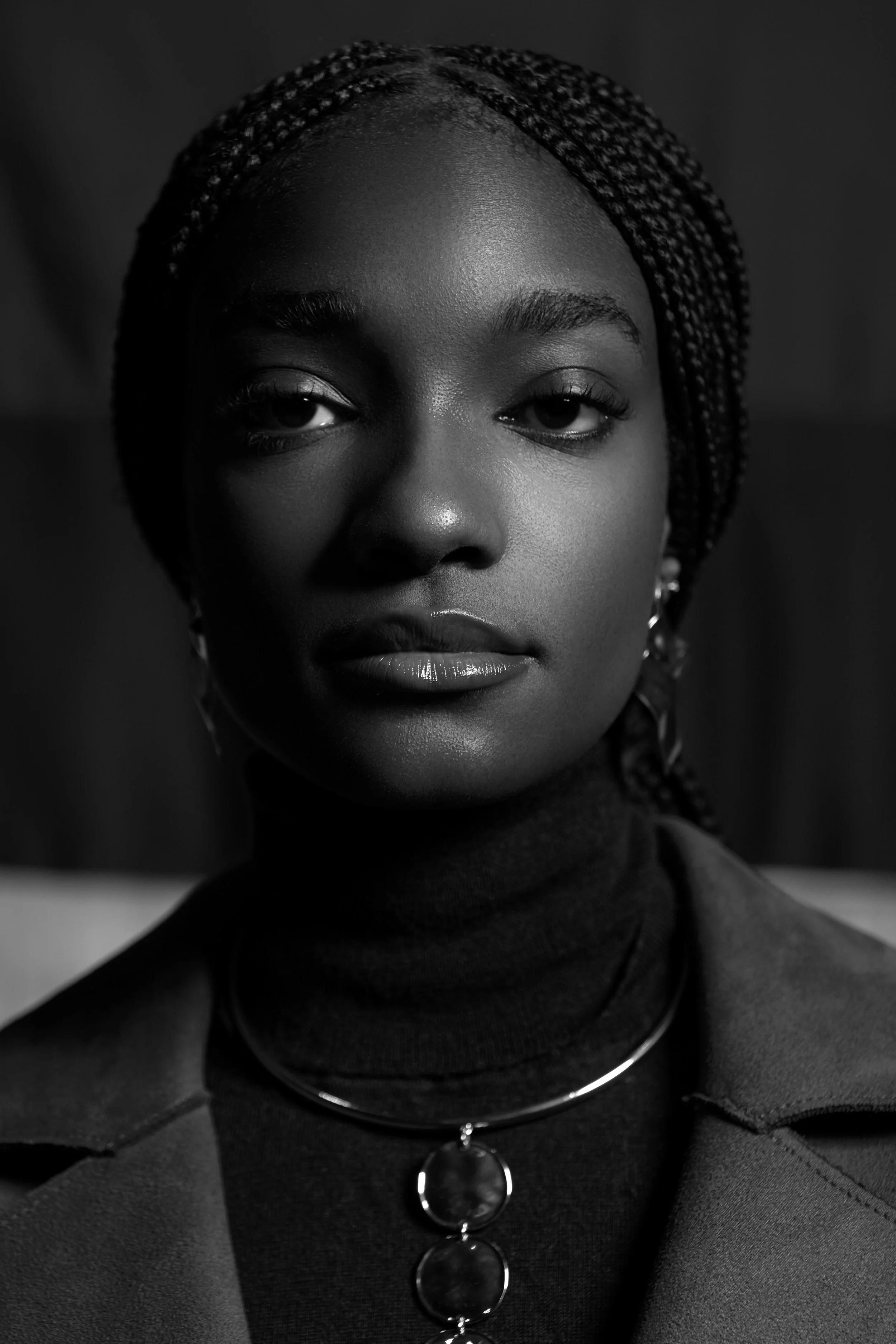 Portrait en noir et blanc d'une femme avec des tresses coiffées en arrière, portant un col noir et un haut à boutons avec des cercles métalliques. Elle a un regard direct.
