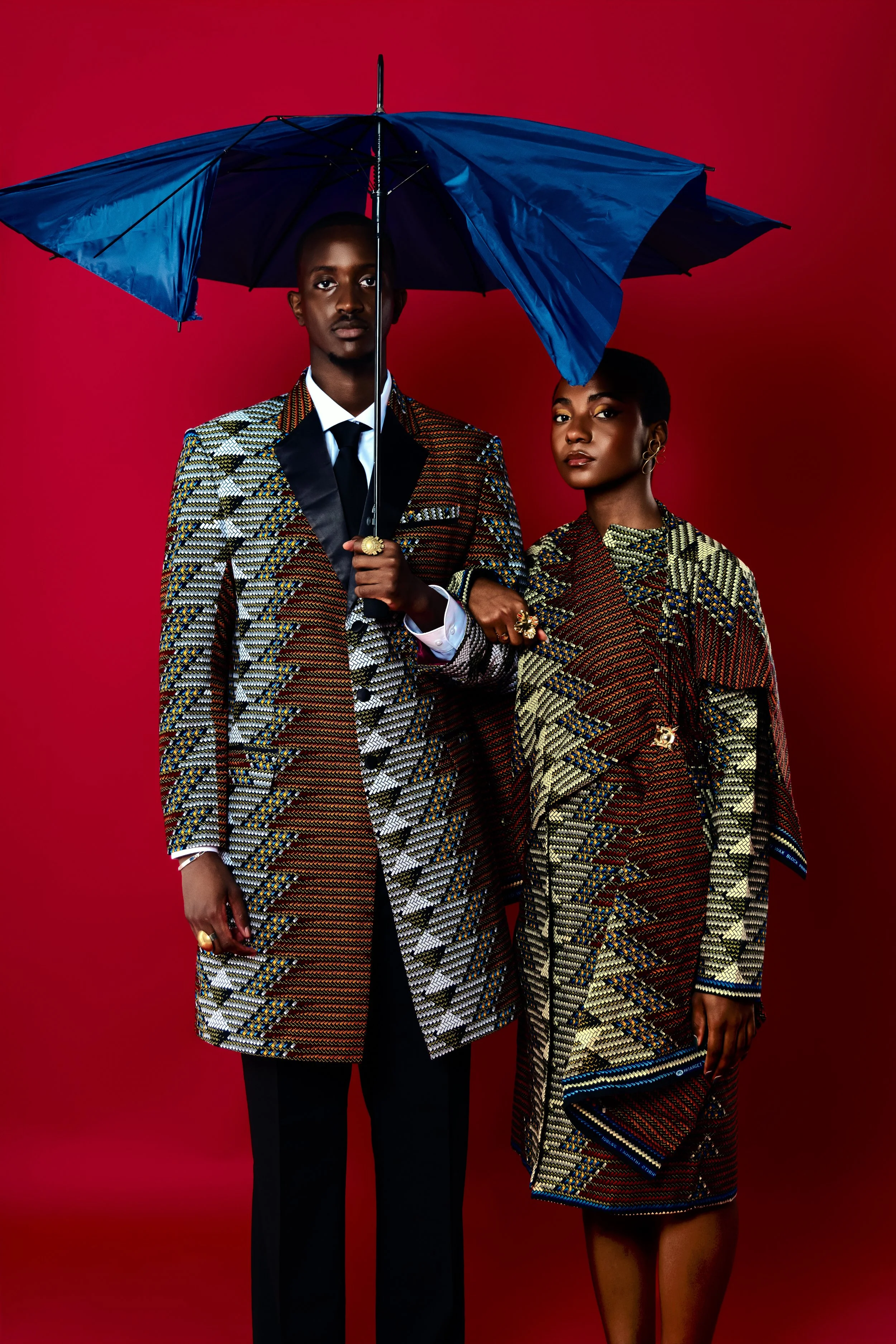Deux personnes habillées en vêtements de mode africaine posent contre un fond rouge, l'une tenant un parapluie bleu.