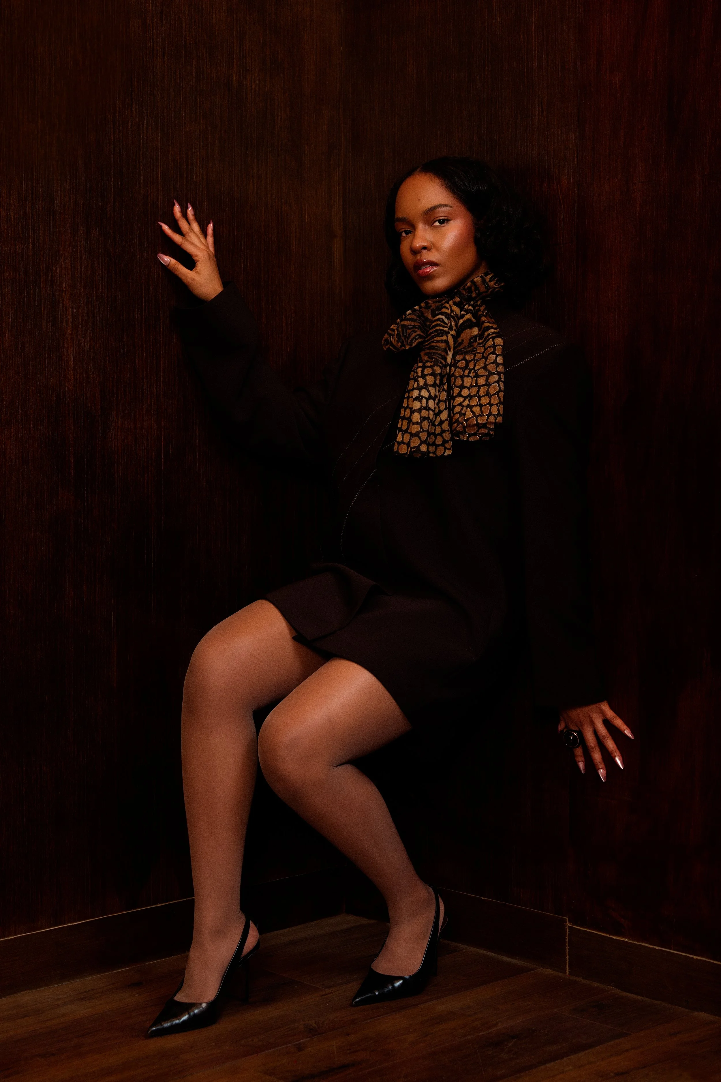 Une femme assise contre un mur en bois, portant une robe noire, des talons noirs, une écharpe à motif léopard et portant un bijou noir à la main.