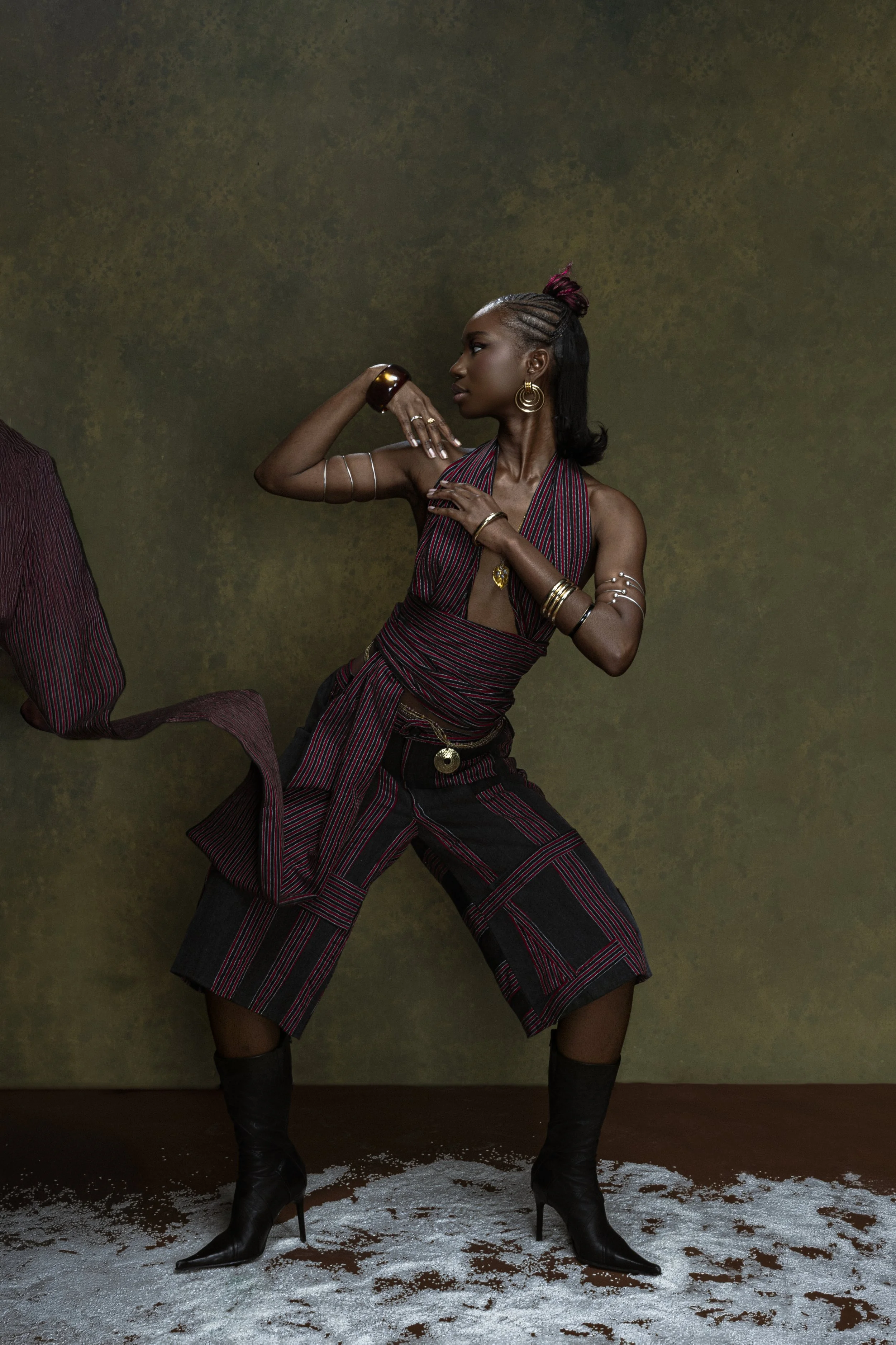 Une femme debout portant un haut à rayures rouges et noires, des accessoires en or, des pantalons à motif à rayures coordonnés, et des bottes à talons hauts, dans une pose artistique contre un fond vert foncé.