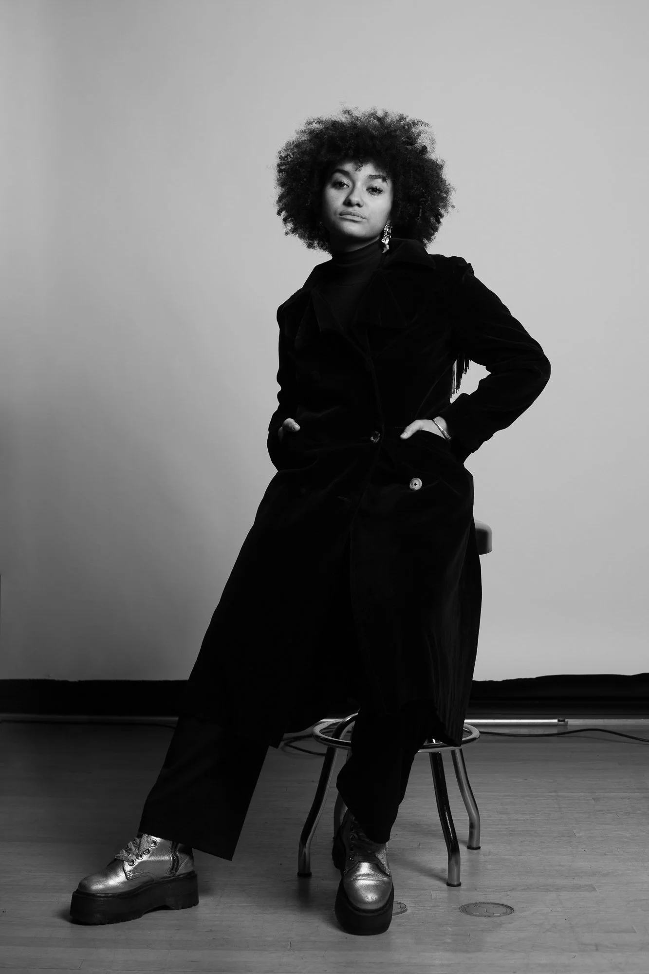 Portrait en noir et blanc d'une femme assise sur une chaise, portant une veste noire, un pantalon large, des chaussures à plateforme argentées, avec de grosses boucles d'oreilles, dans un studio avec un fond gris clair.