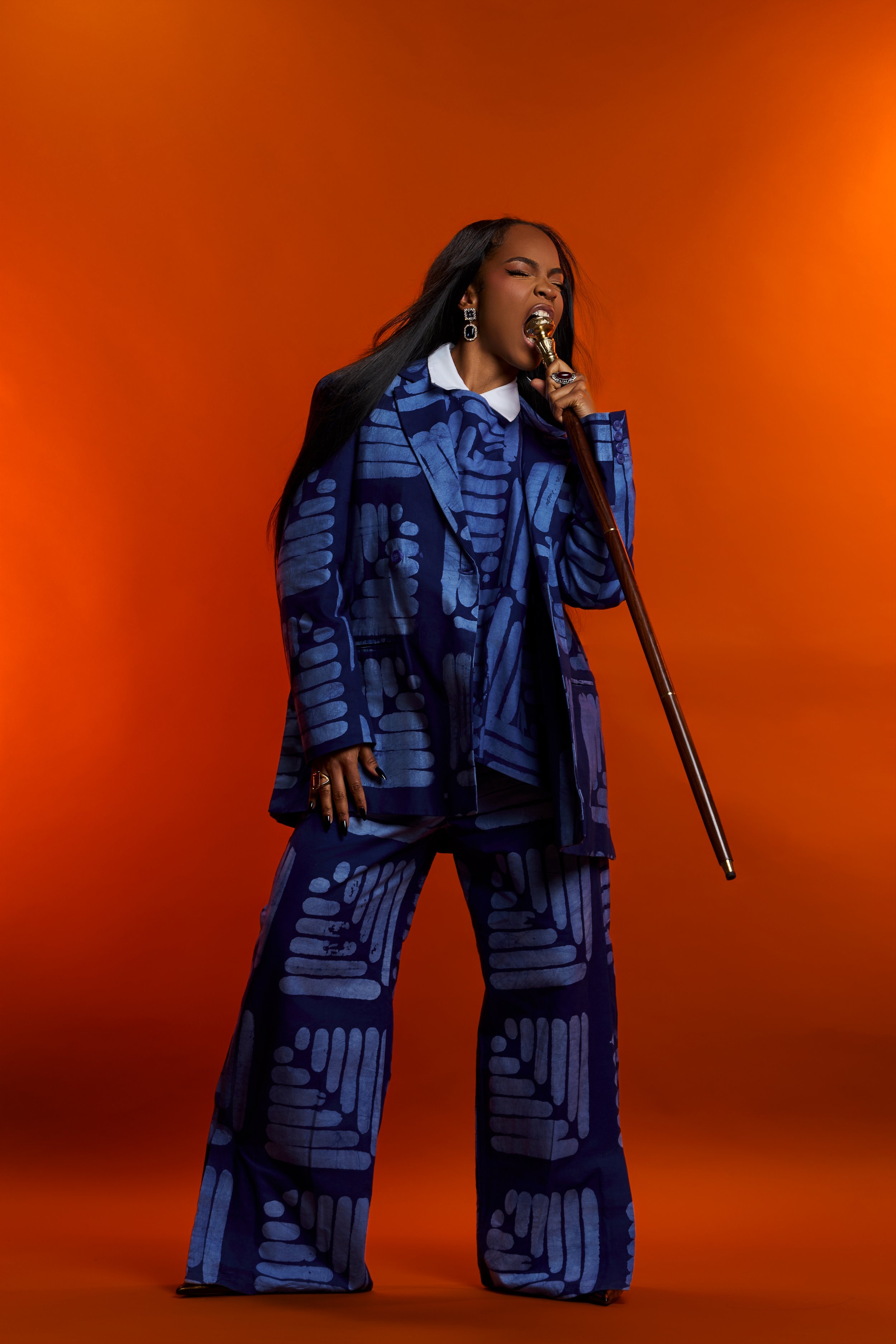 Une femme noire avec de longs cheveux noirs, portant un costume à motifs bleus, pose de côté tout en chantant dans un microphone en or, contre un fond orange.