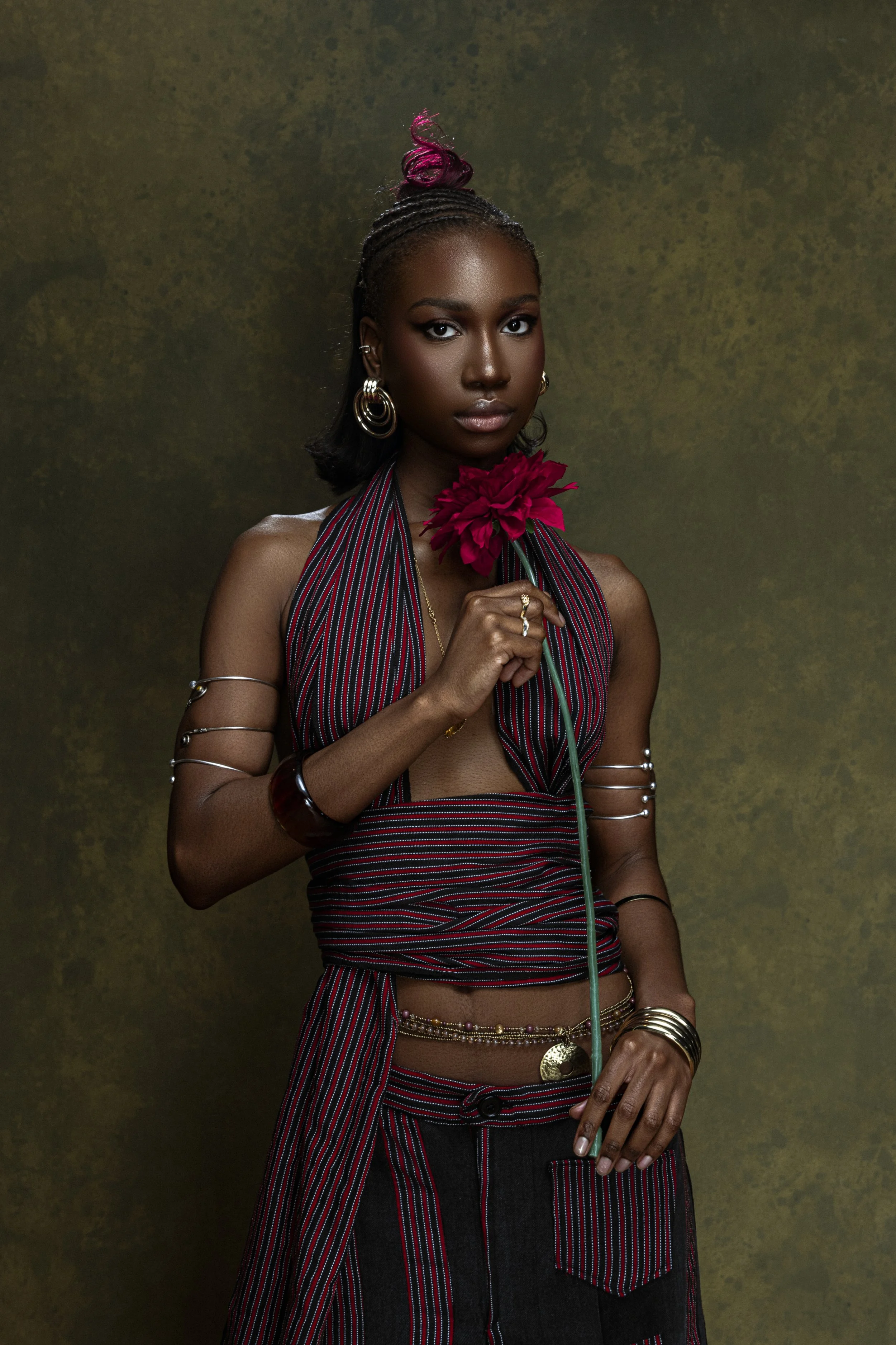 Une femme noire avec des cheveux noirs et roses, portant un haut à rayures rouges et noires, tenant une fleur rouge à la main, avec des bijoux or et argent, devant un fond vert foncé.