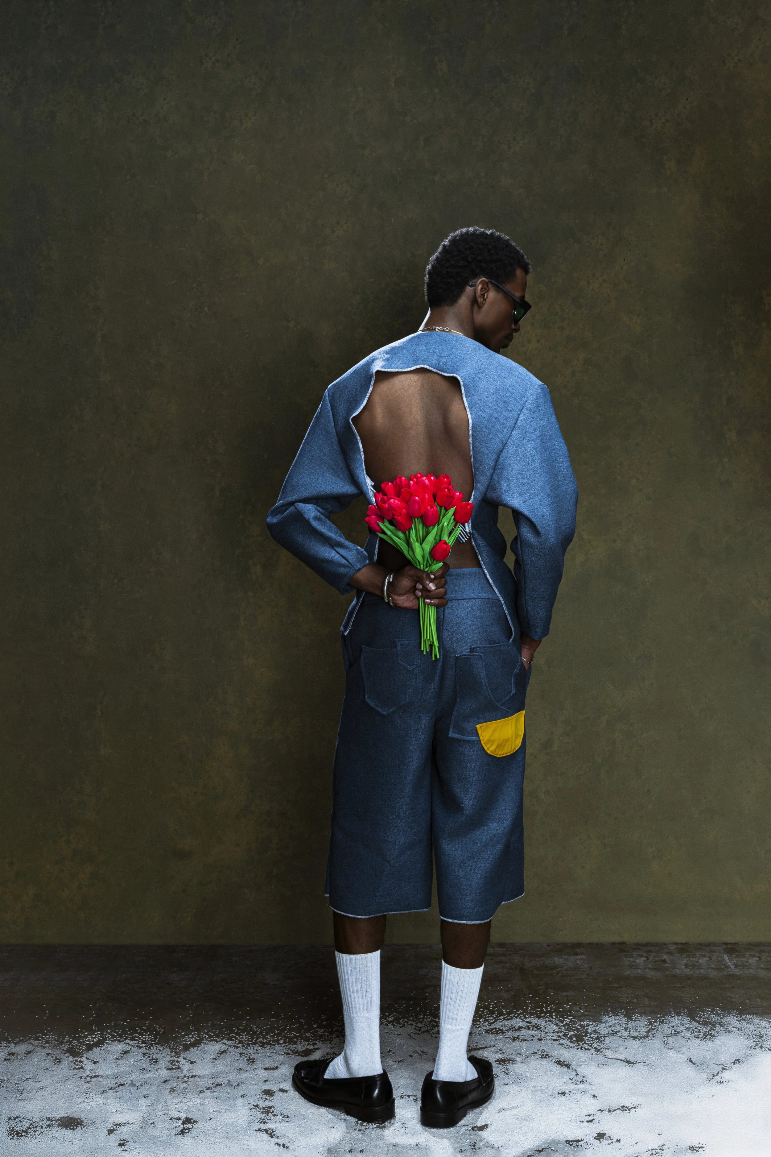 Une personne vue de dos, portant une veste en jean avec un dos ouvert, tenant un bouquet de tulipes rouges derrière le dos, portant un pantalon en denim, des chaussettes blanches jusqu'au genou, et des chaussures noires, debout devant un mur sombre.