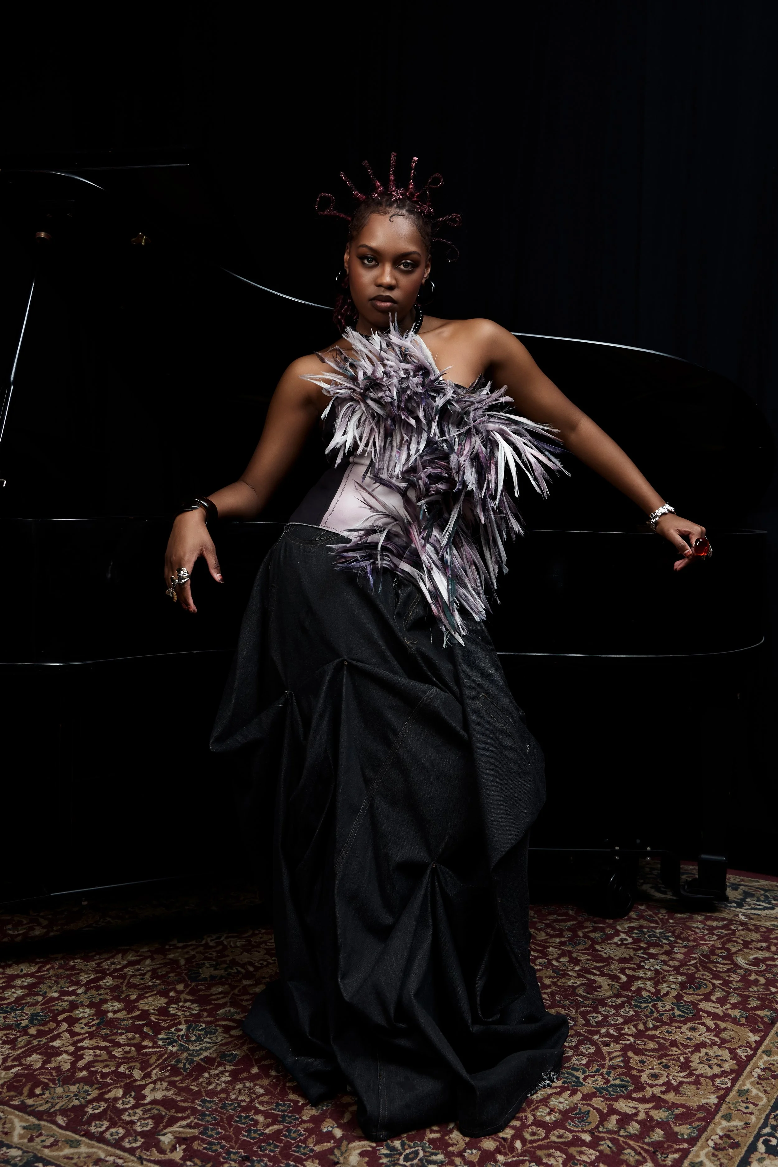 Une femme noire pose devant un piano noir dans une pièce sombre, portant une jupe longue noire et un haut orné de plumes violettes, avec des bijoux et un chignon décoré de fils violets.