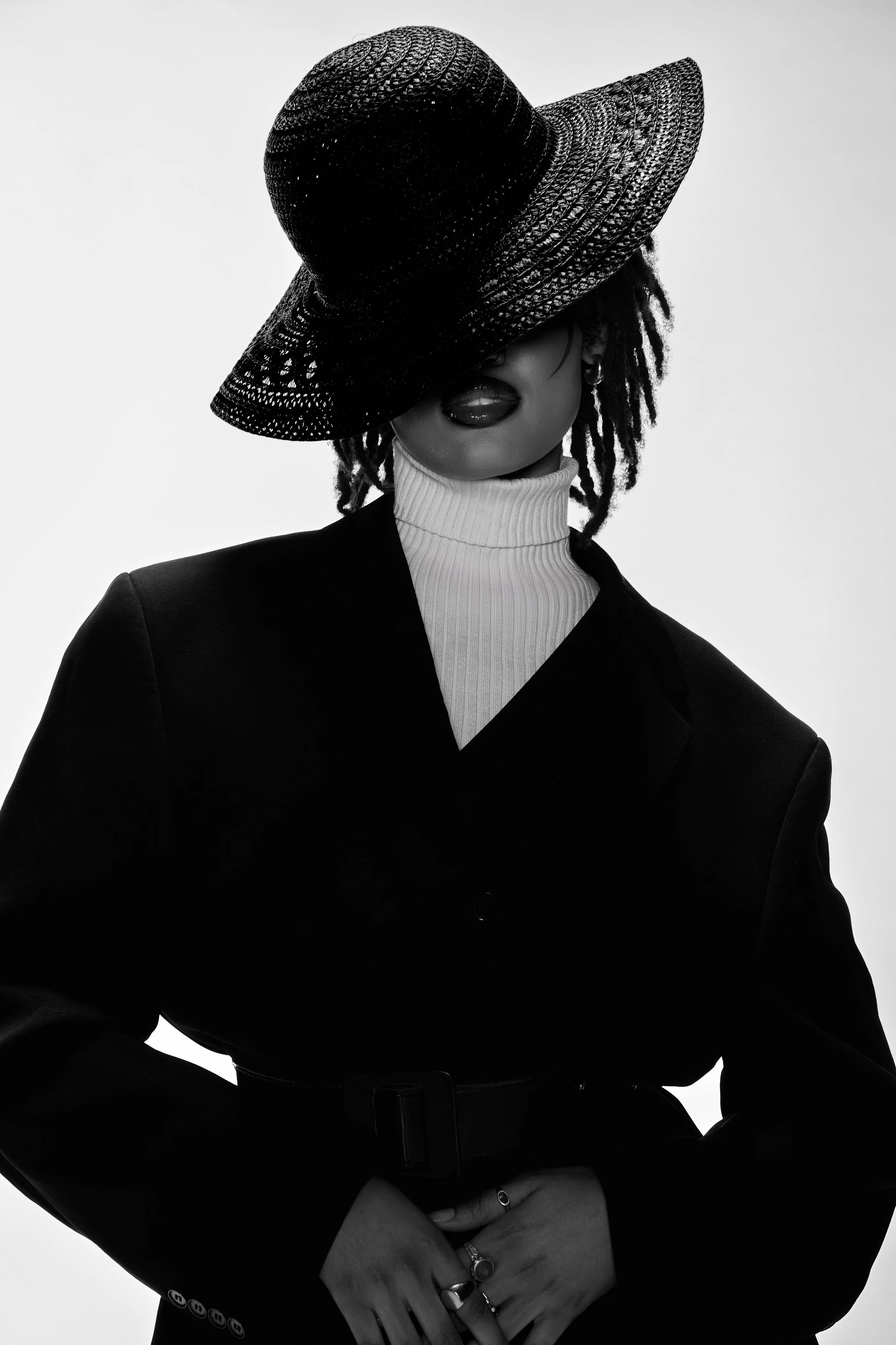 Femme portant un chapeau noir en paille, vêtue d'un manteau noir et d'un col en tresse blanche, avec plusieurs bagues, dont une en forme de cercle.
