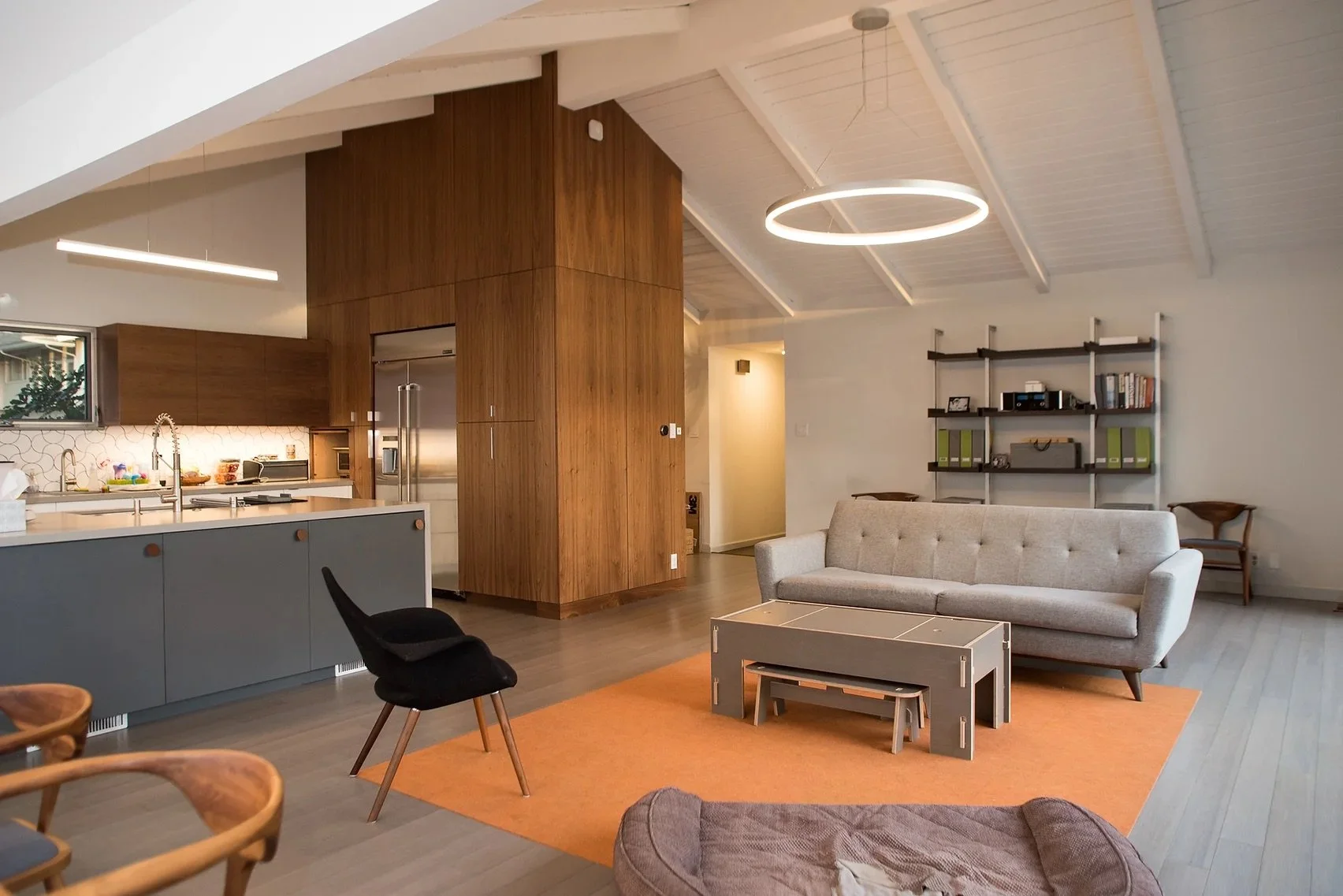 Modern living room with kitchen, featuring a gray sofa, orange rug, wooden cabinets, pendant lights, and shelving with books.